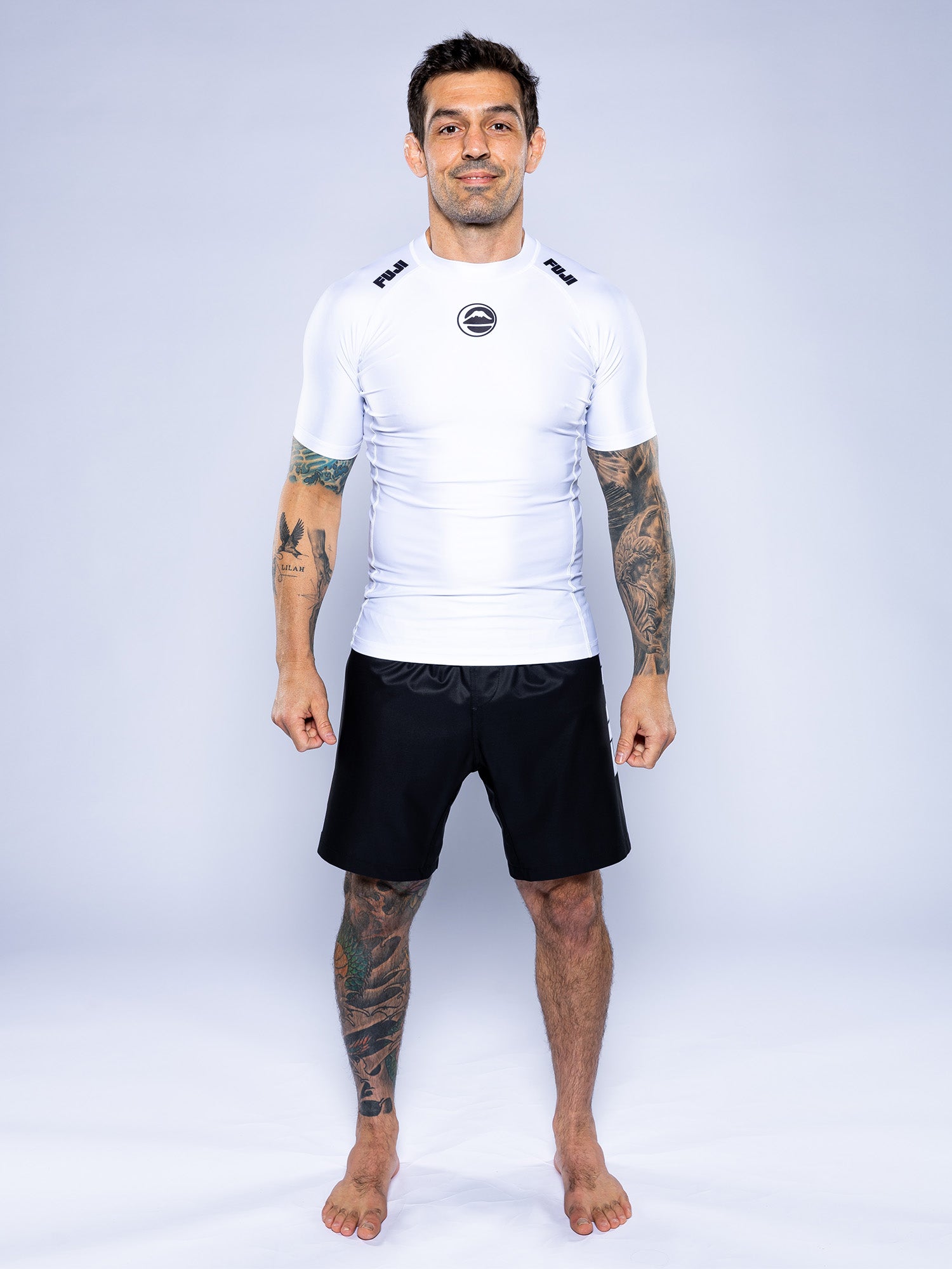 A tattooed man stands barefoot on a white background, wearing the Baseline 2.0 Short Sleeve Rashguard White and black shorts, facing forward with a slight smile.