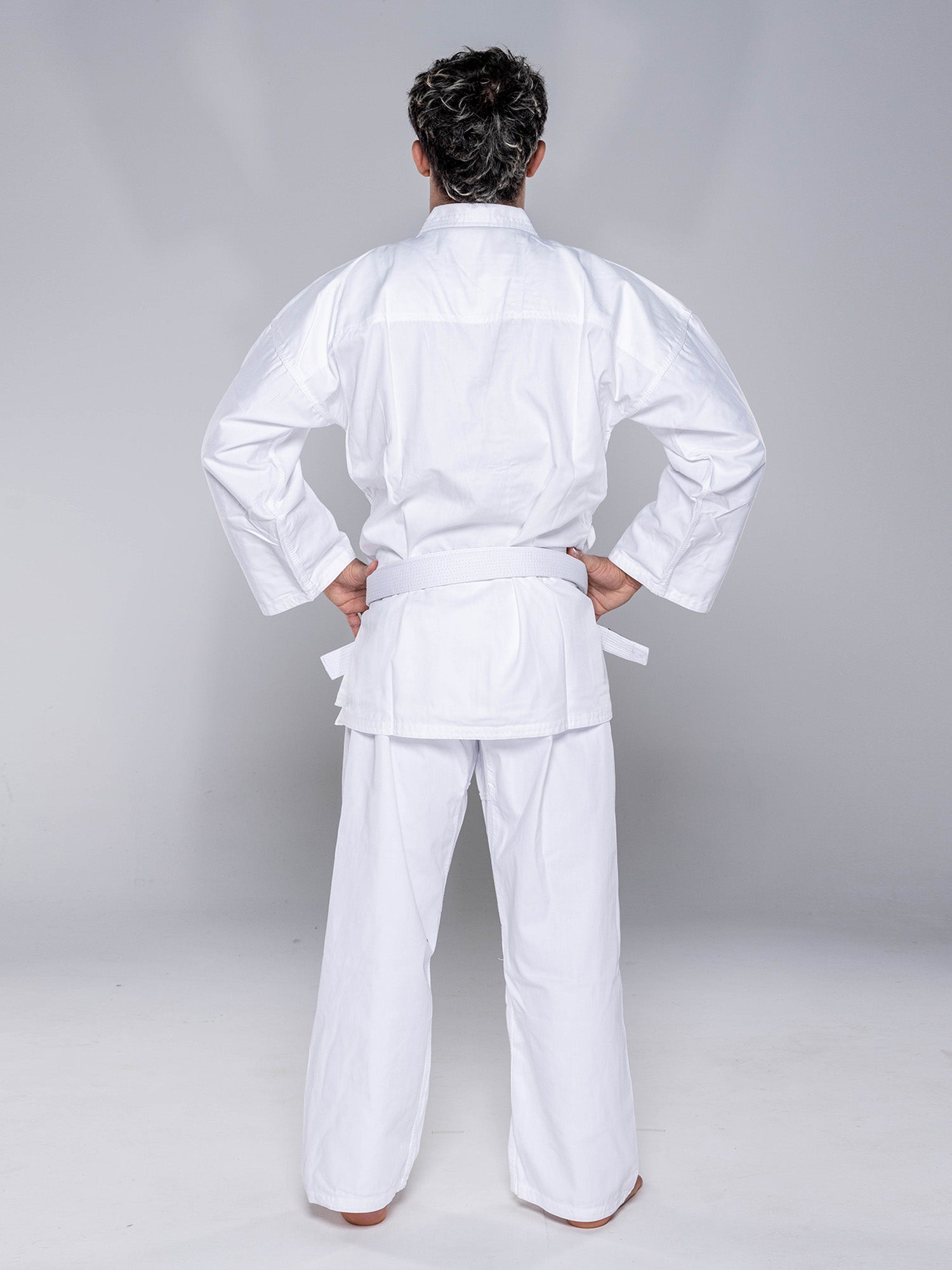 A person in a Keiko Karate Gi - Starter with belt White stands barefoot, hands on hips, facing away from the camera against a plain light background.