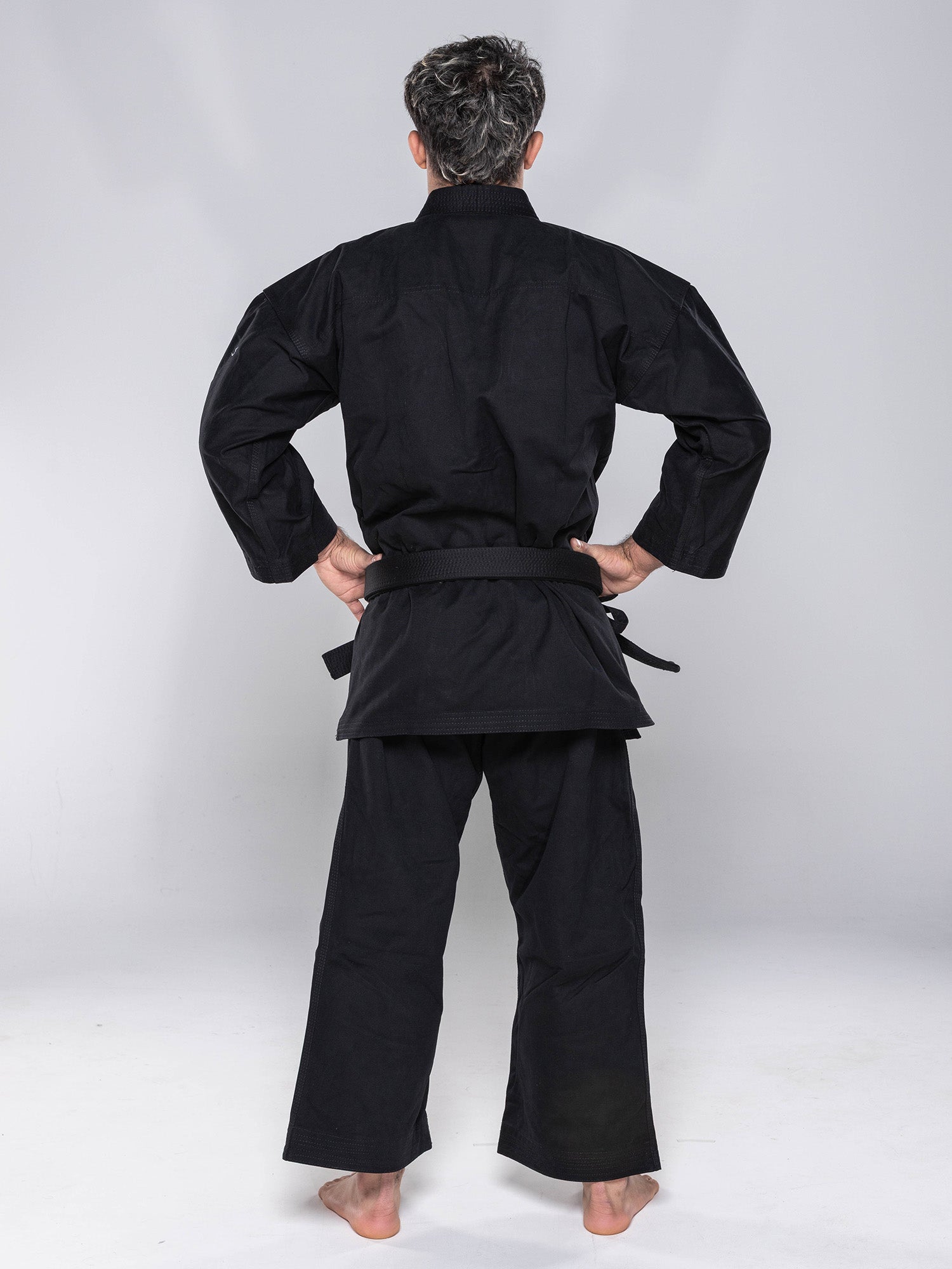 A person stands barefoot with hands on hips, facing away from the camera, wearing the Kaizen Karate Gi Advanced Black and black belt in a plain, light-colored studio setting.