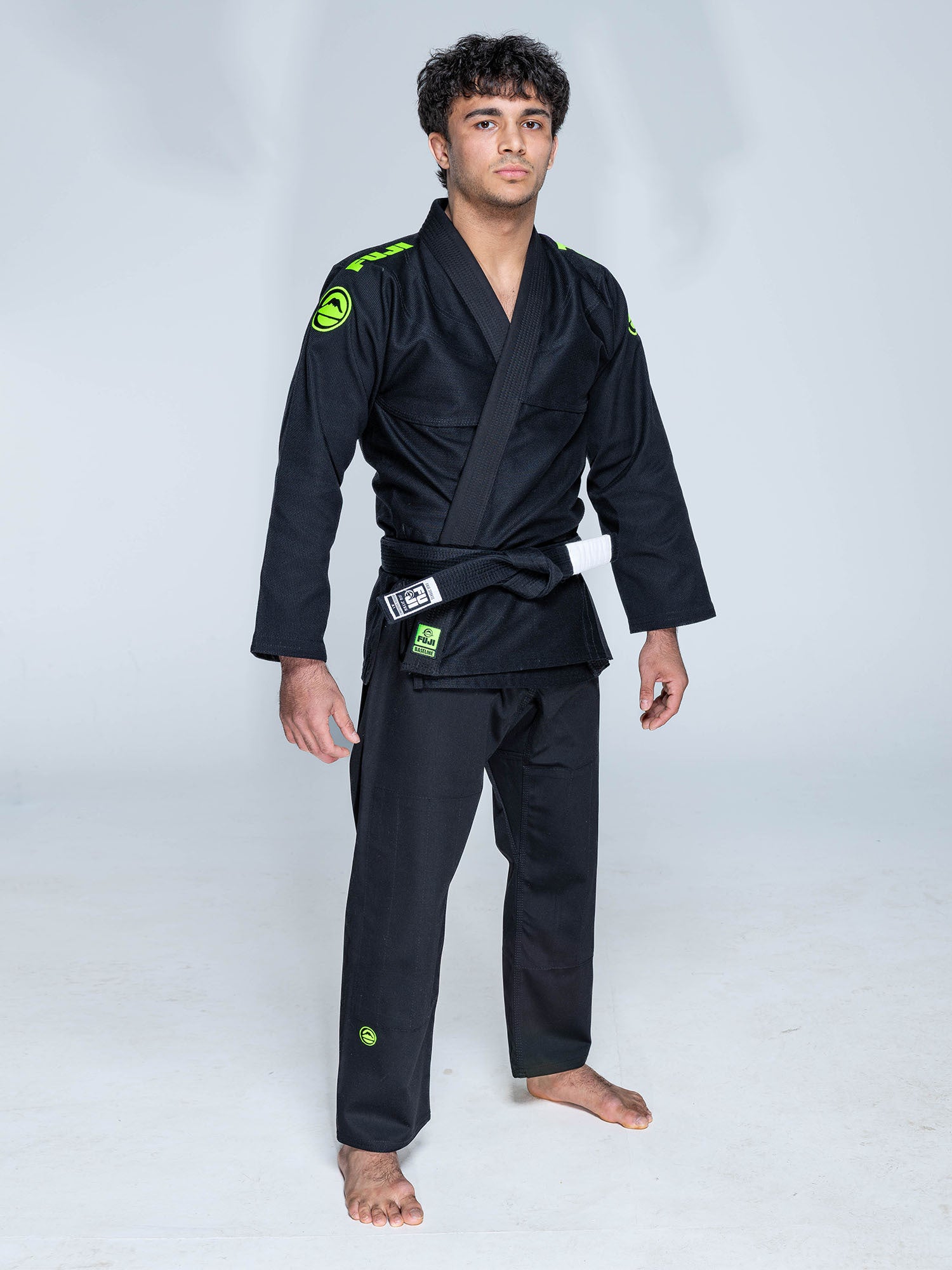 A young man stands barefoot, facing forward against a plain light background, wearing the Baseline 2.0 Slim Fit BJJ Gi Black Green with green logos and a black belt.