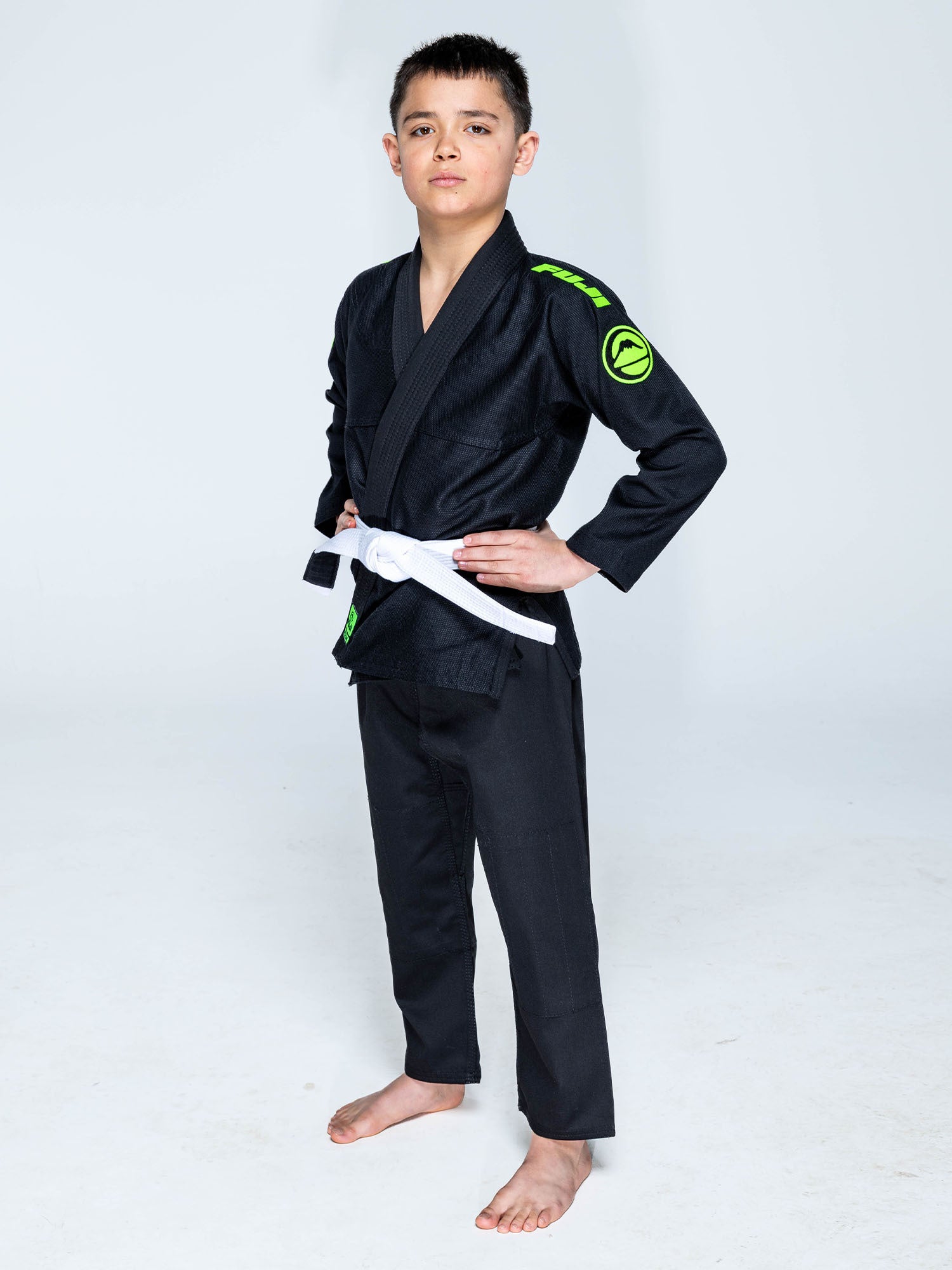 A young boy stands barefoot on a white background, wearing the Kids Baseline 2.0 Slim Fit BJJ Gi in Black Green, complete with a white belt at his waist. He rests his hands on his hips and looks confidently at the camera.