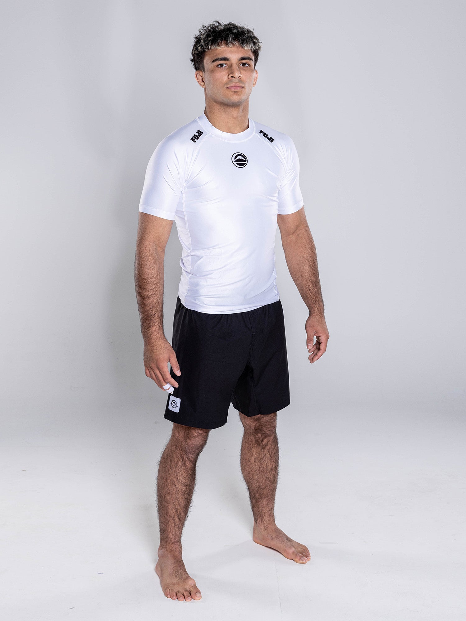 A man with short curly hair stands barefoot on a white background, wearing the Baseline 2.0 Short Sleeve Rashguard White with black logos and black athletic shorts, arms relaxed at his sides and a neutral expression.
