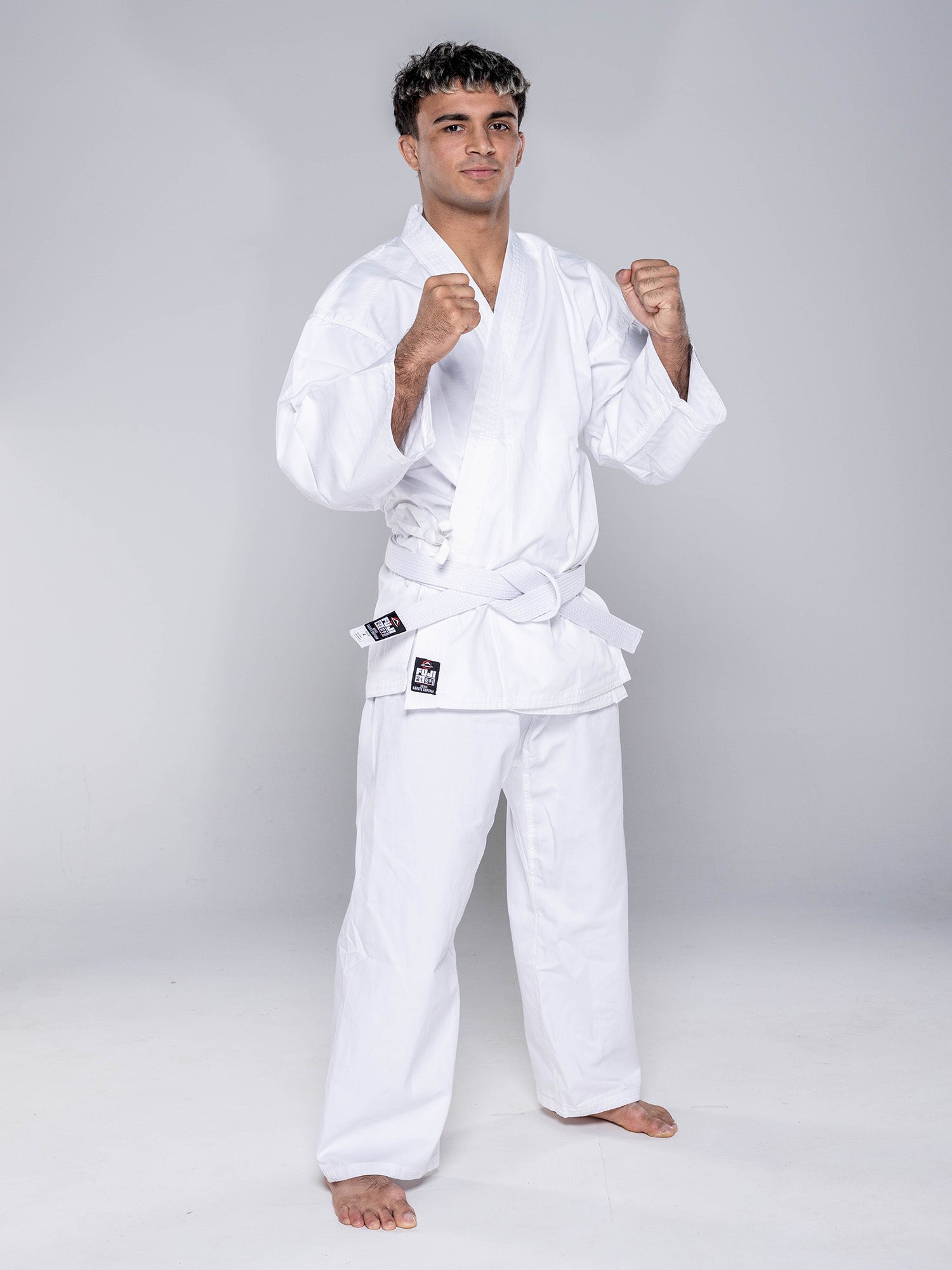 A young man stands barefoot in a fighting stance, fists raised, confidently posing in the Keiko Karate Gi - Starter with belt White against a plain gray background.