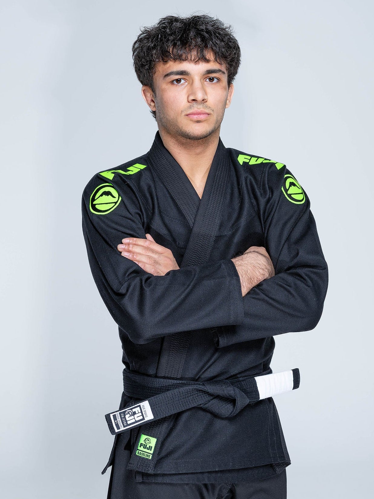 A young man with curly hair stands with arms crossed, wearing the Baseline 2.0 Slim Fit BJJ Gi Black Green and a black belt, against a plain light gray background.
