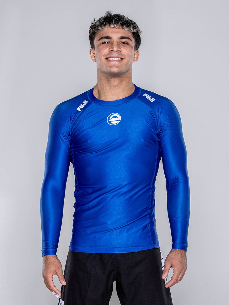 A young man stands barefoot on a white floor, smiling. He wears the Baseline 2.0 Long Sleeve Rashguard Navy and black shorts against a plain, light gray background.