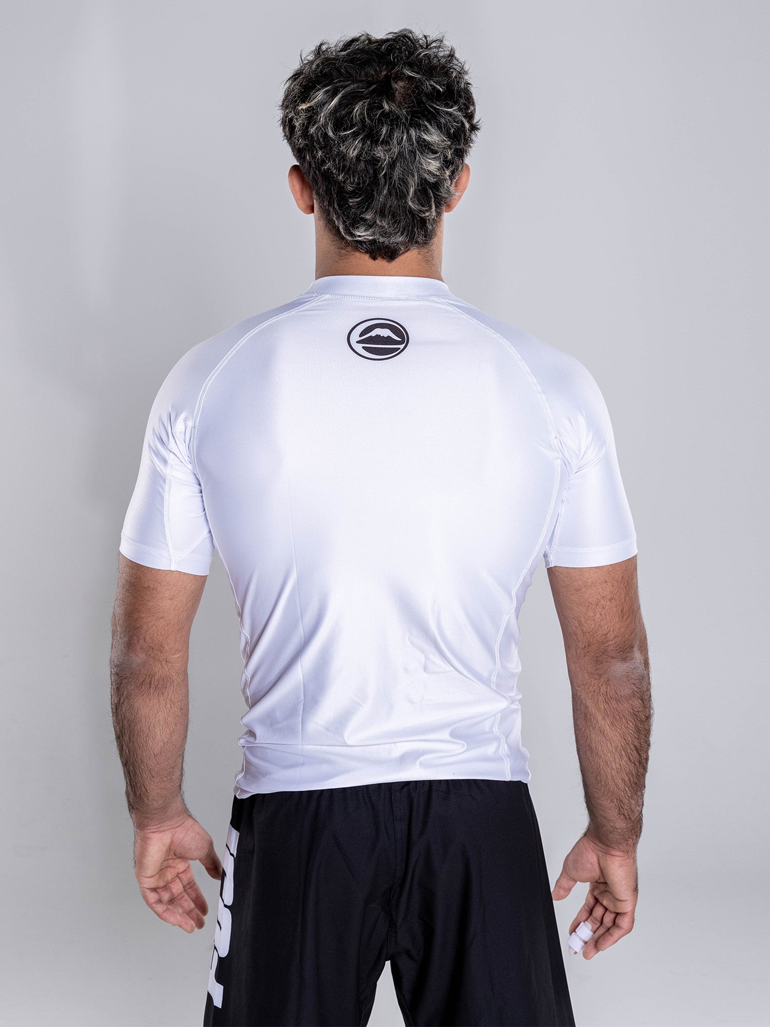 A man with short, dark hair and subtle highlights stands with his back to the camera, wearing the Baseline 2.0 Short Sleeve Rashguard White featuring a circular logo on the upper back, paired with black sports shorts against a plain light gray background.