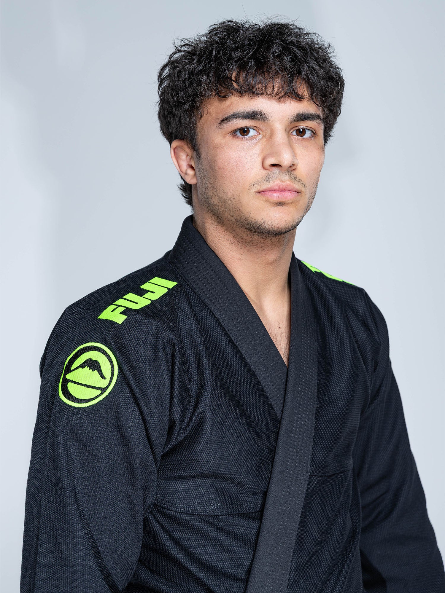 A young man with curly hair wears the Baseline 2.0 Slim Fit BJJ Gi in black and green, featuring Fuji logos on the shoulders, as he stands calmly in front of a plain background.