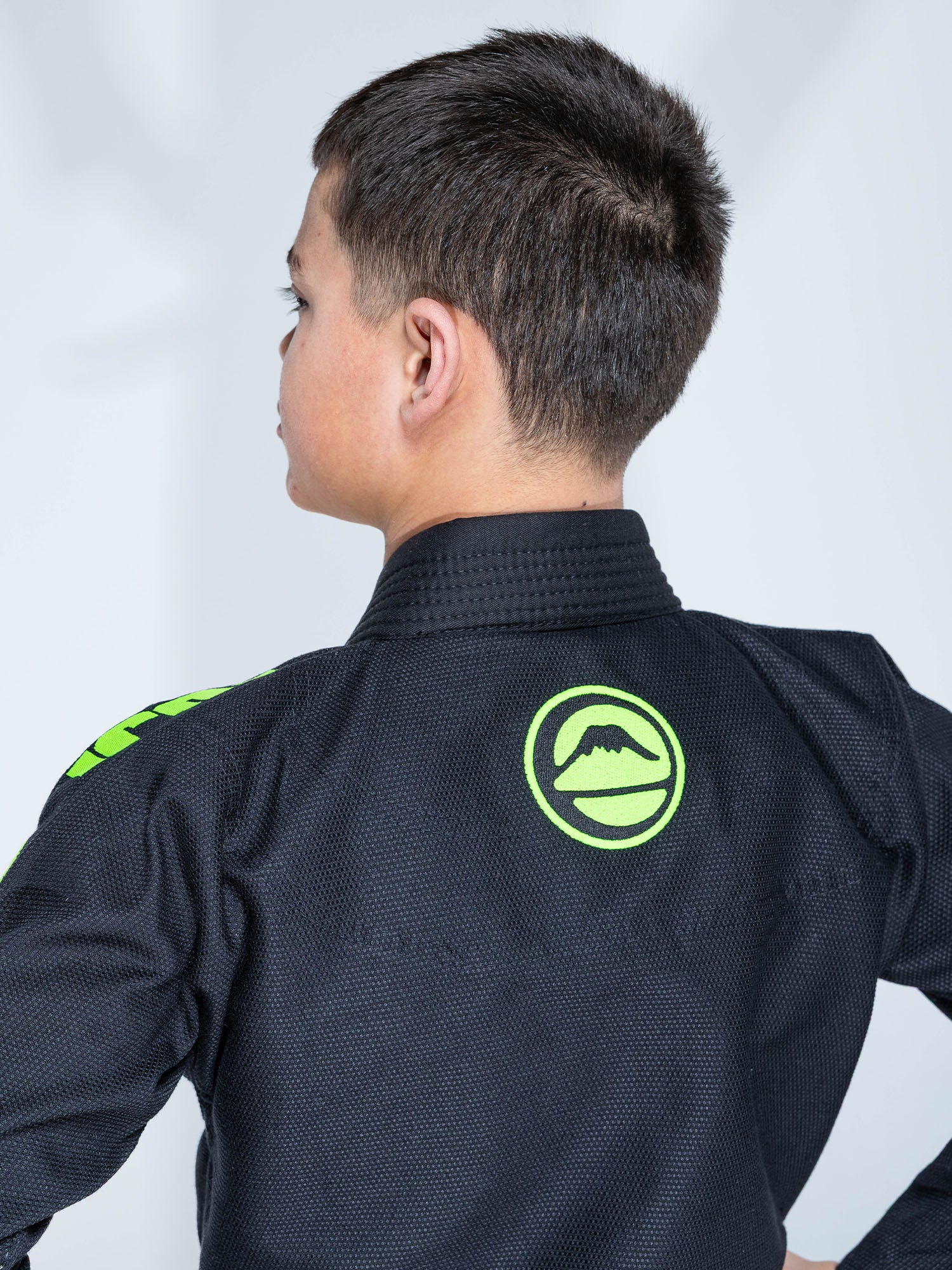 A young boy with short brown hair wears the Kids Baseline 2.0 Slim Fit BJJ Gi Black Green, featuring a green mountain logo on the back, shown from behind against a plain light background with his profile visible.