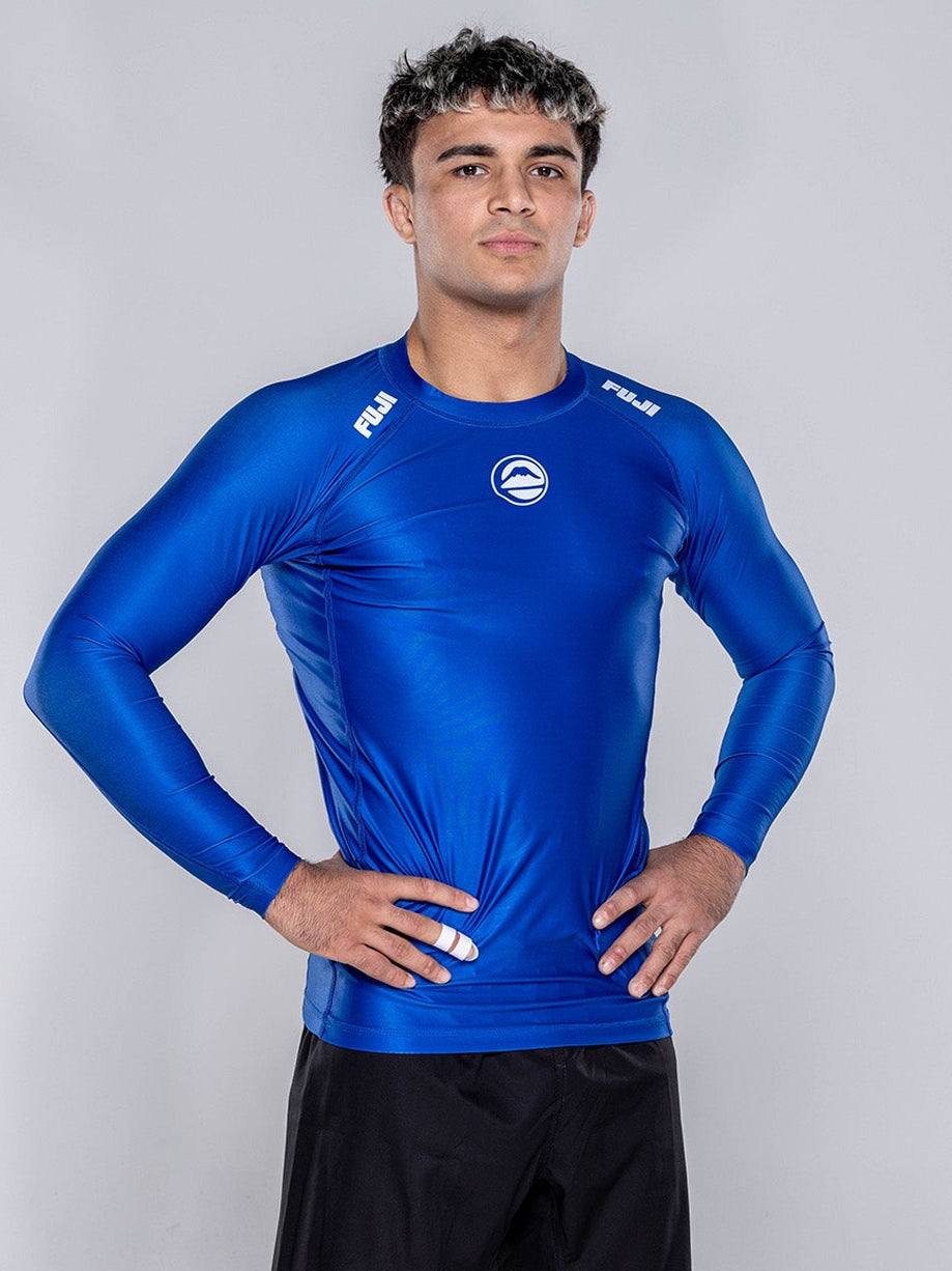 A young man stands barefoot on a white background, confidently wearing the Baseline 2.0 Long Sleeve Rashguard Navy and black shorts with his hands on his hips.