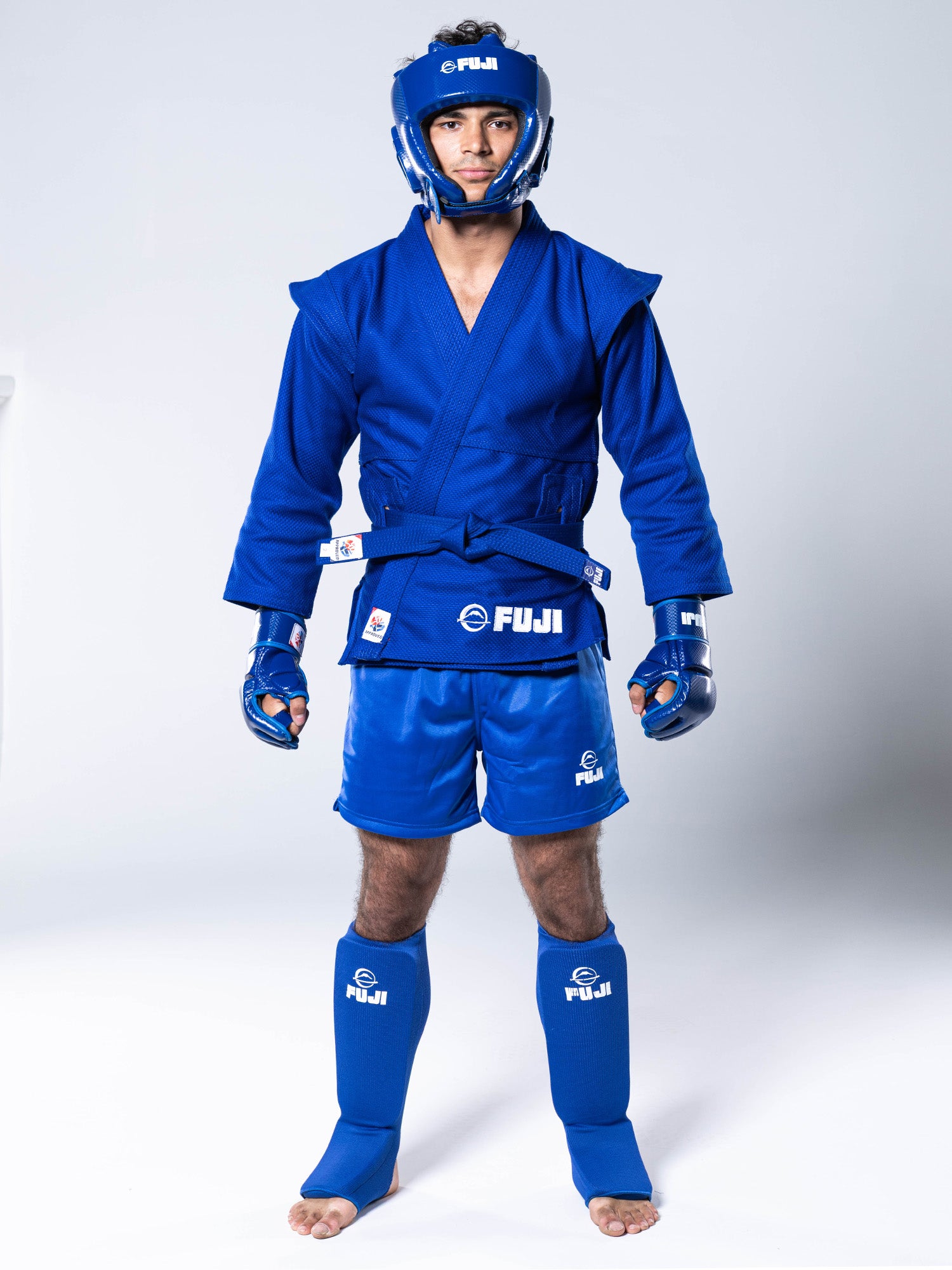 A person stands facing forward on a plain white background, wearing FUJI-branded blue martial arts gear that includes the FIAS Sambo Combat Headgear Blue, matching gloves, gi top, shorts, shin guards, and other protective equipment.