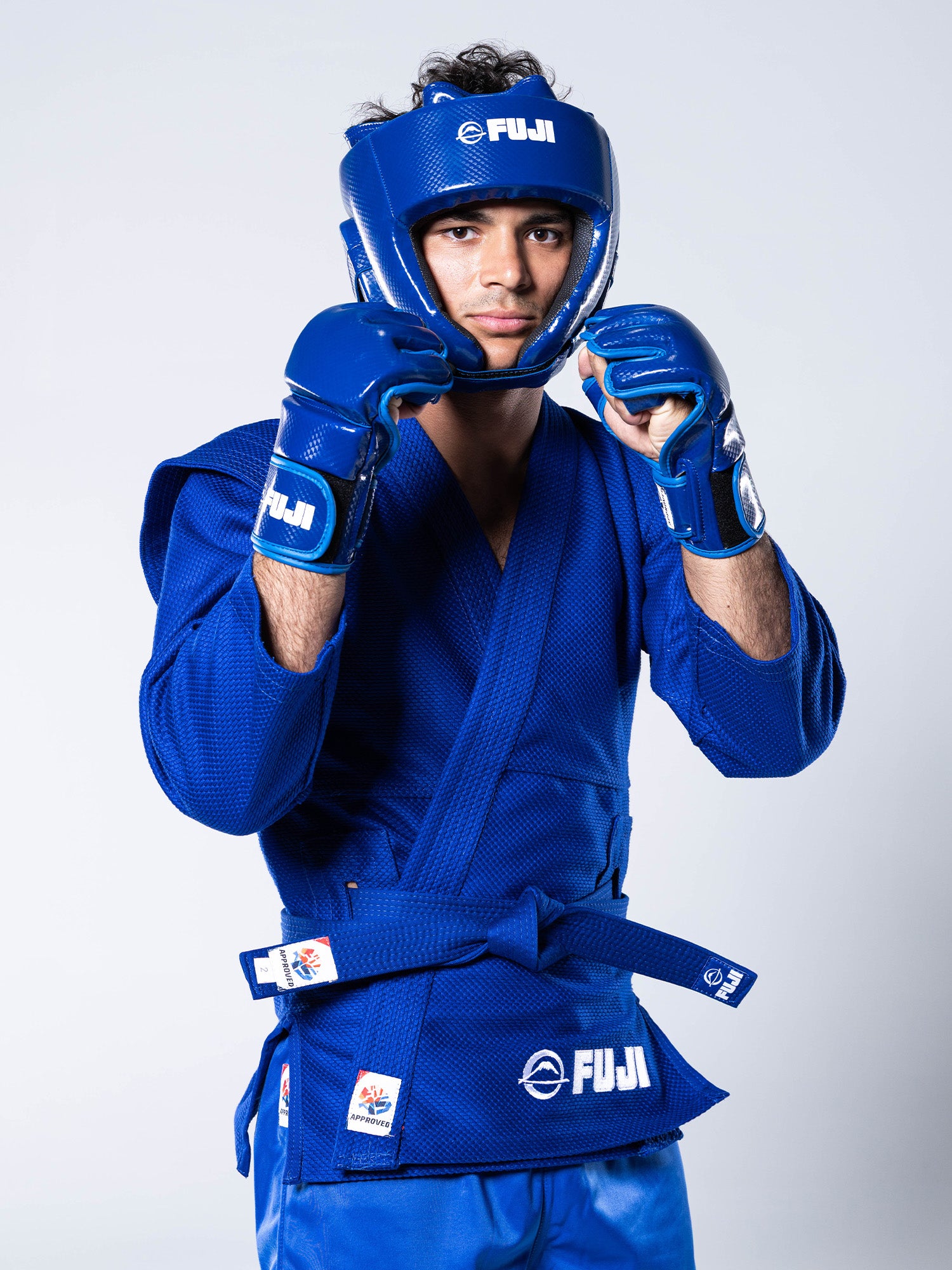A martial artist wearing the FIAS Sambo Combat Headgear Blue, dressed in matching blue protective gear and gi with gloves, stands in a fighting stance against a plain background.
