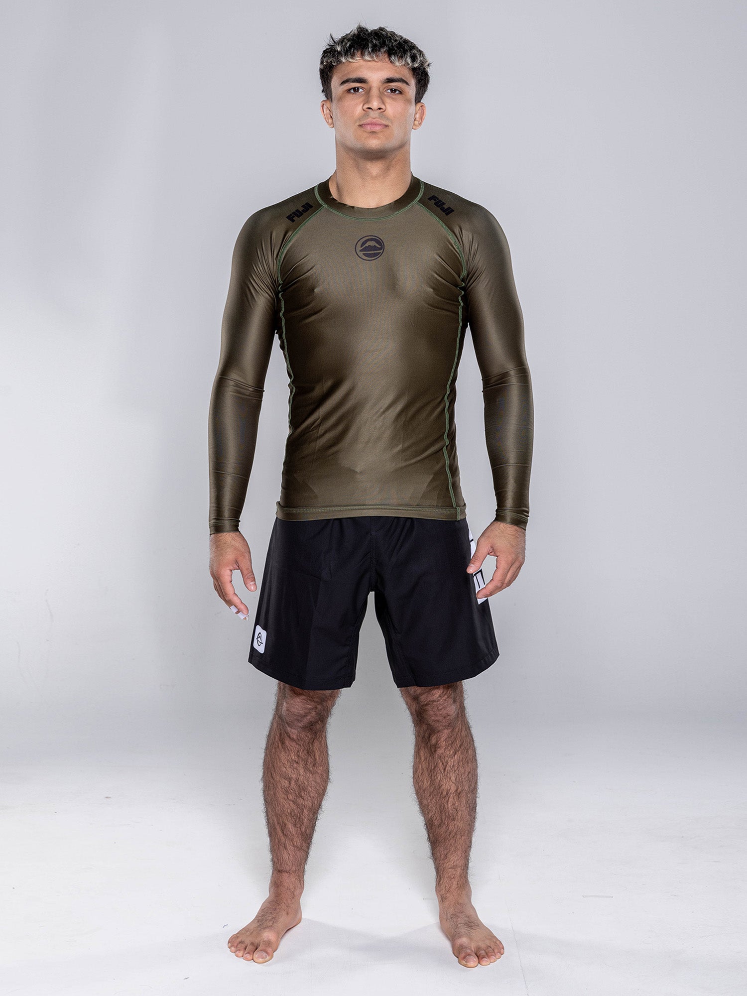 A man stands barefoot on a white floor, wearing the Baseline 2.0 Long Sleeve Rashguard in Military Green and black athletic shorts, facing forward with arms relaxed by his sides against a plain light gray background.