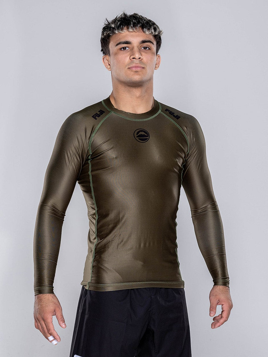 A person stands barefoot on a white floor, wearing the Baseline 2.0 Long Sleeve Rashguard in Military Green and black shorts. The background is plain light gray, and the person faces forward with a neutral expression.