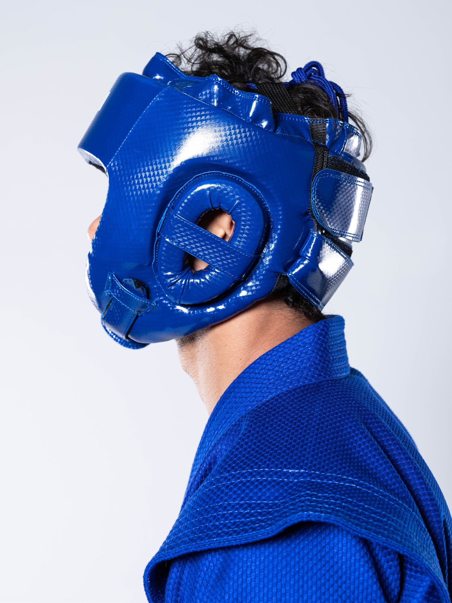 Profile view of a person wearing the FIAS Sambo Combat Headgear Blue and a matching blue textured martial arts uniform against a plain white background.