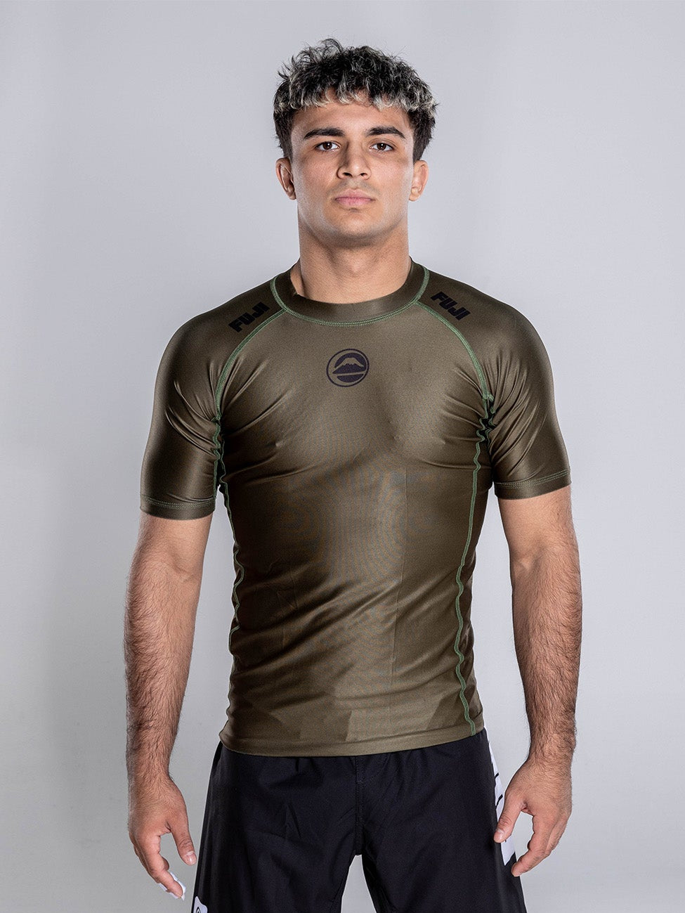 A man stands barefoot on a white background, wearing the Baseline 2.0 Short Sleeve Rashguard in Military Green and black shorts, facing forward with his arms relaxed at his sides.