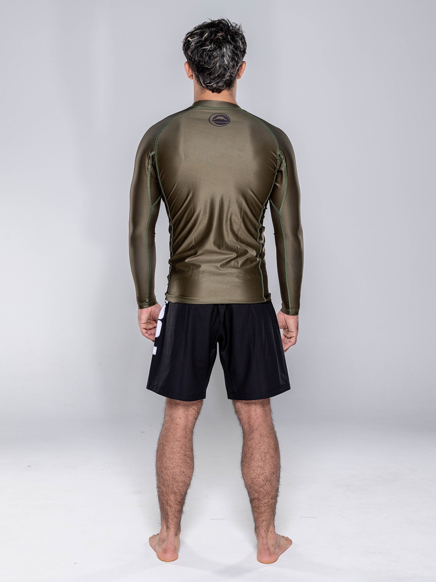 A man stands barefoot on a white floor, facing away from the camera. He is wearing the Baseline 2.0 Long Sleeve Rashguard in Military Green and black shorts, with a plain light gray background.