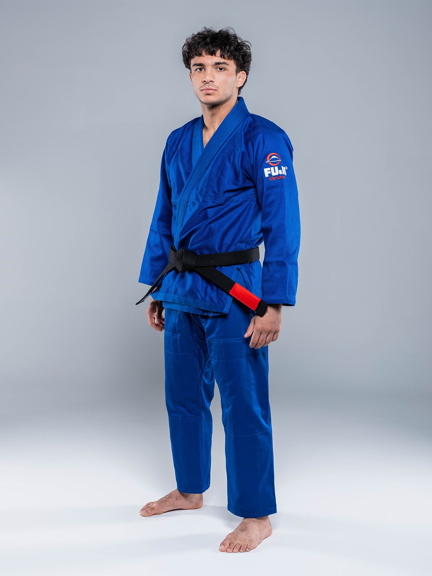 A man stands barefoot on a plain background, facing forward with a neutral expression, wearing the Lightweight BJJ Gi Blue with a black belt.