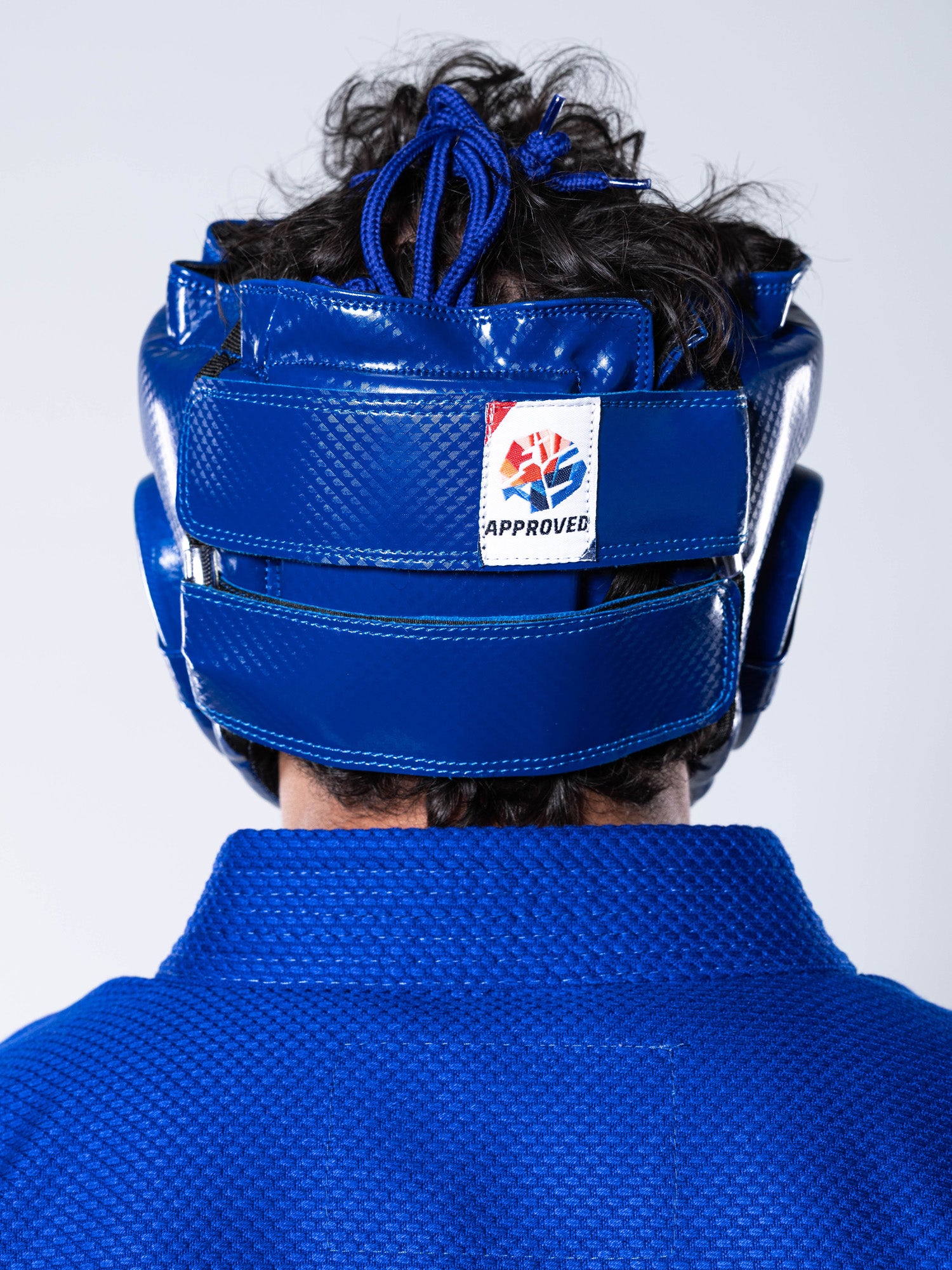 A person is shown from behind wearing a FIAS Sambo Combat Headgear Blue and a blue martial arts gi; the helmet features a logo and the word “APPROVED” on a white patch.