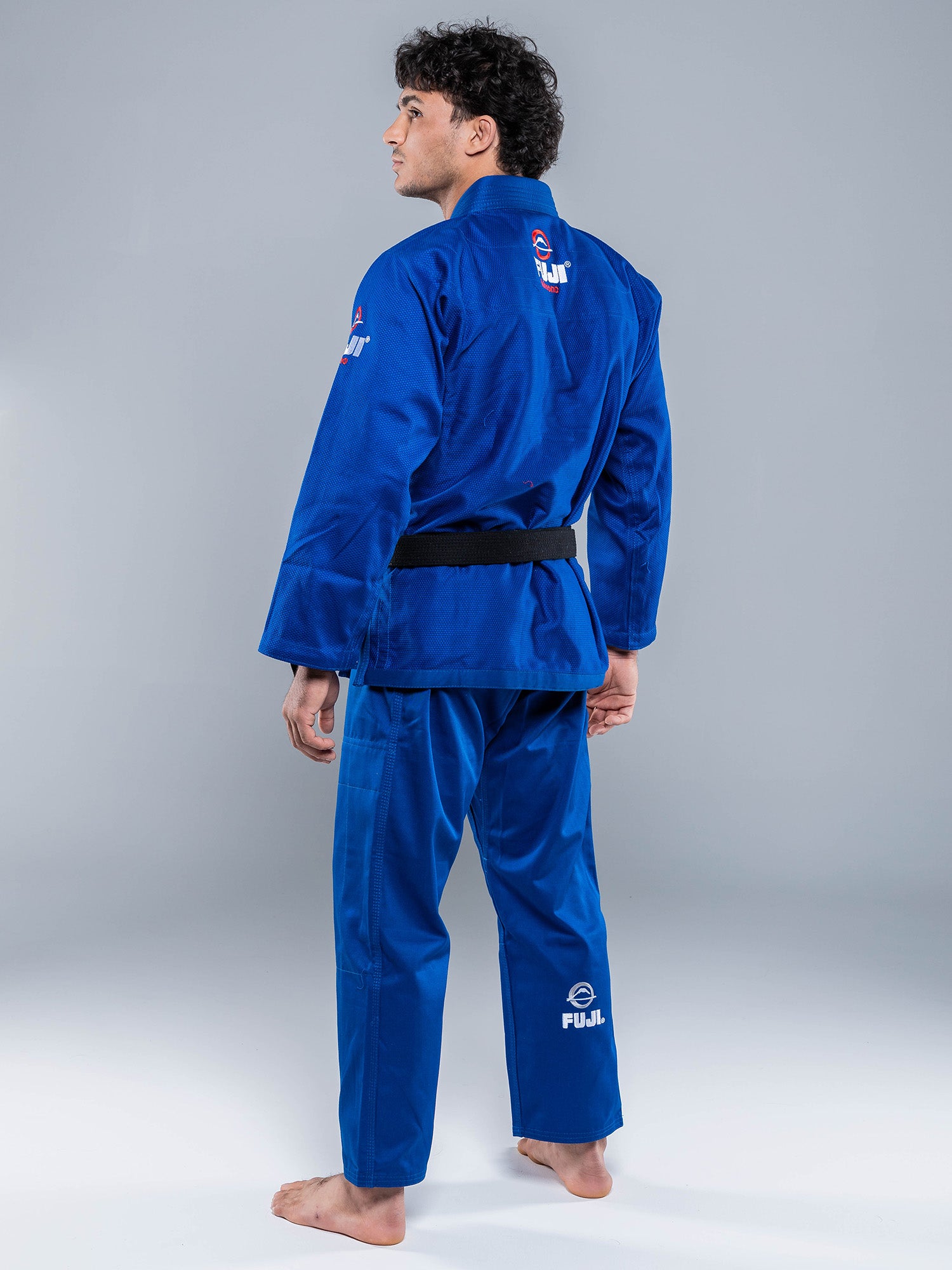 A person wearing the Lightweight BJJ Gi Blue with a black belt stands barefoot on a light surface, facing away and slightly sideways. The gi displays white logos on the back, arm, and pant leg.