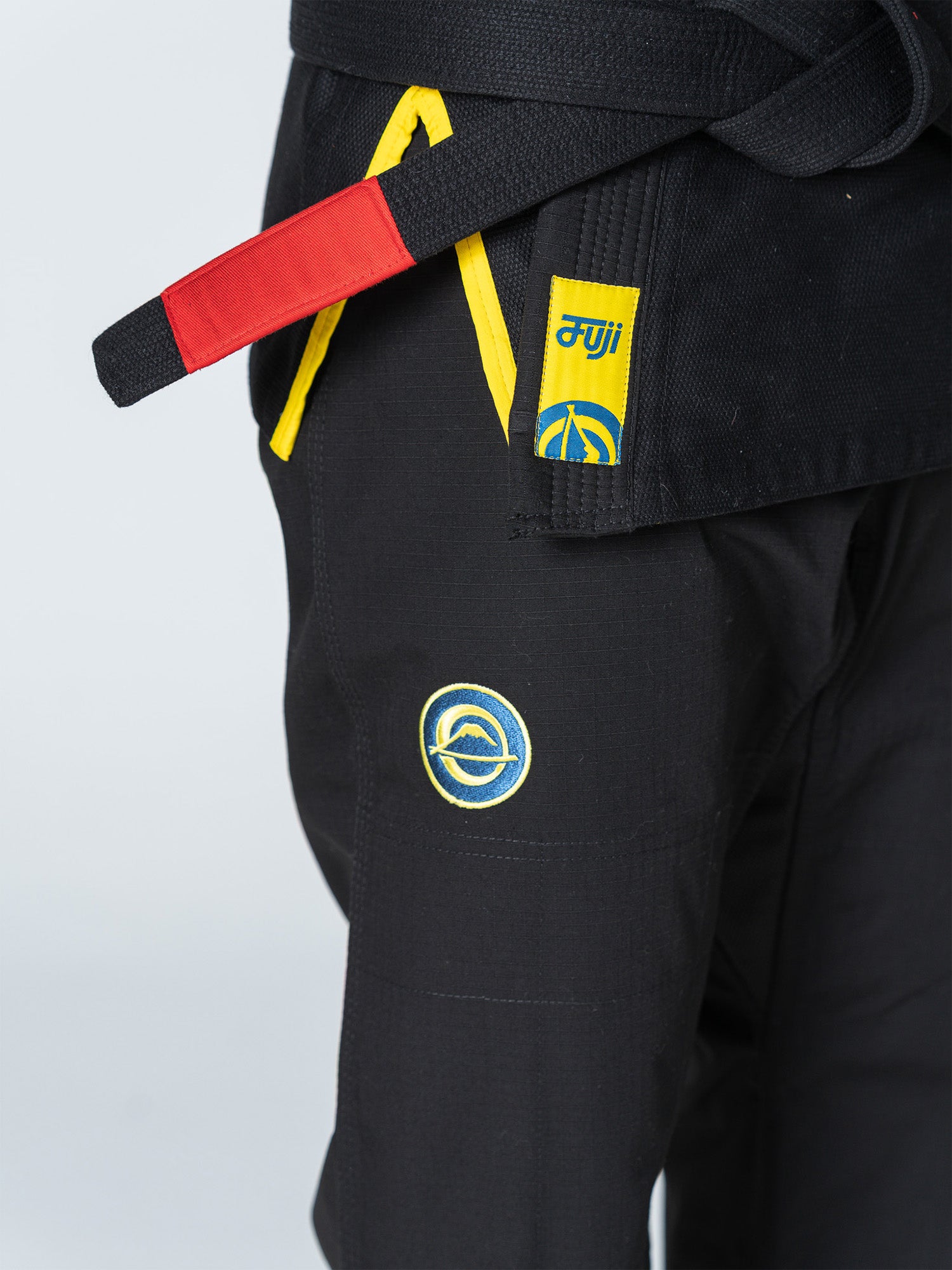 A close-up of a person wearing the Special Edition Holiday Flow Tech Jiu Jitsu Gi Black, featuring yellow-trimmed patches, a black belt with a red bar, and embroidered circular logos on the pants and jacket.