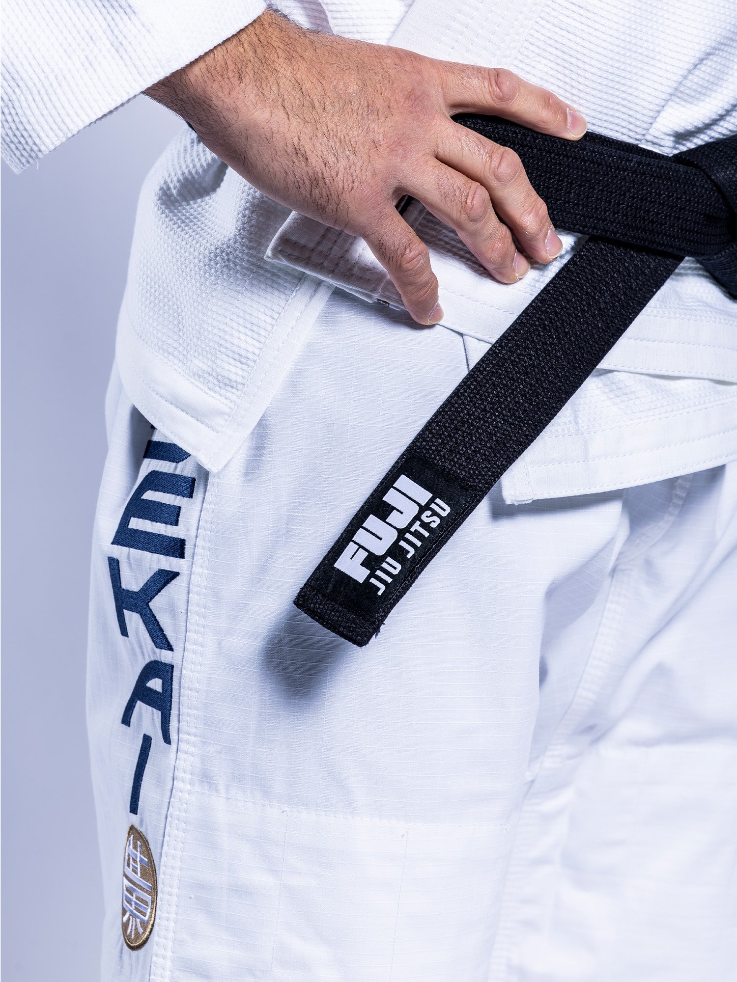 A person wears the SEKAI 3.0 BJJ Gi White Navy and a Fuji Jiu-Jitsu black belt, their hand resting near the belt against a plain background.