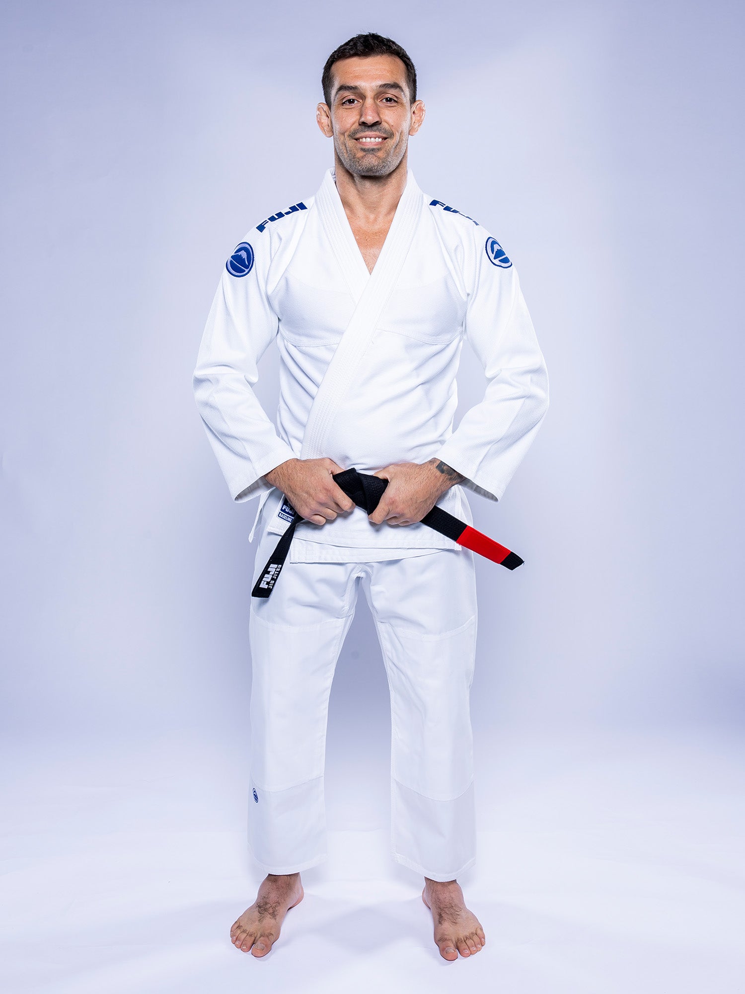A man wearing the Baseline 2.0 BJJ Gi White Navy stands barefoot against a light background, holding a black belt with a red bar and smiling at the camera.