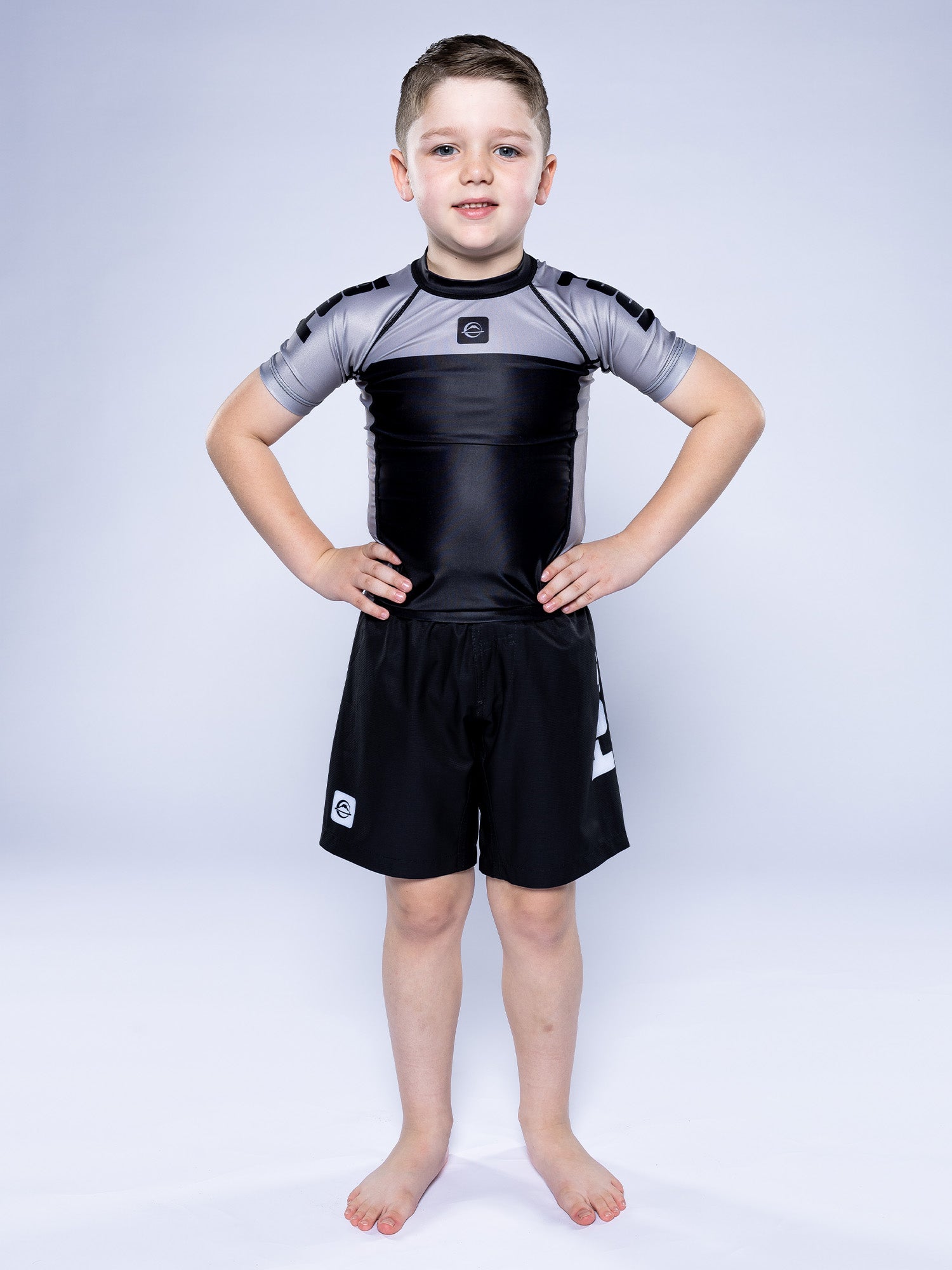 A young boy stands barefoot on a white background, wearing the Kids Competition Ranked Rashguard Grey and black shorts. He faces forward with his hands on his hips and a slight smile.