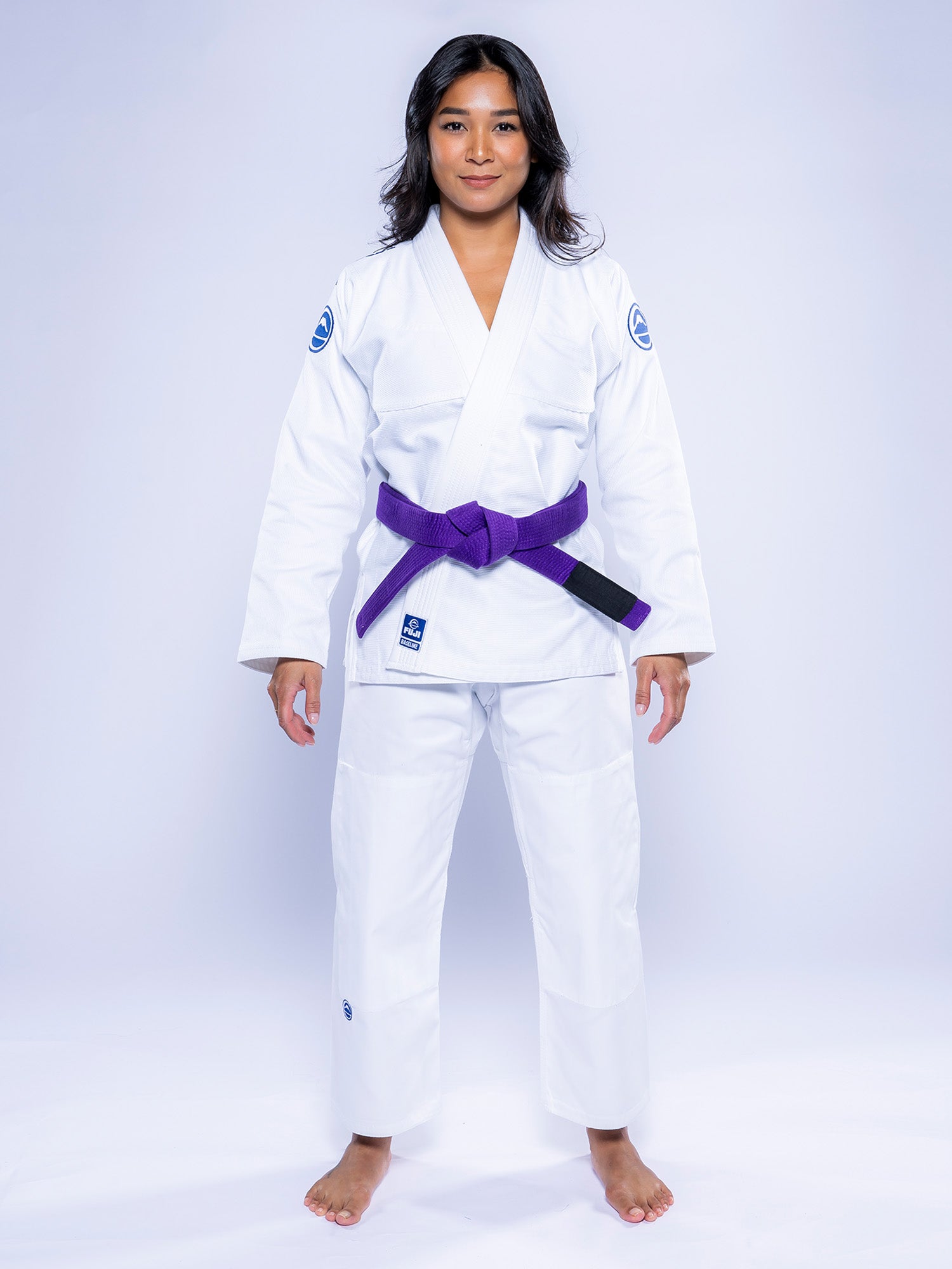 A person stands barefoot on a white background, wearing the Womens Baseline 2.0 BJJ Gi in White Navy with a purple belt, looking forward with a neutral expression.