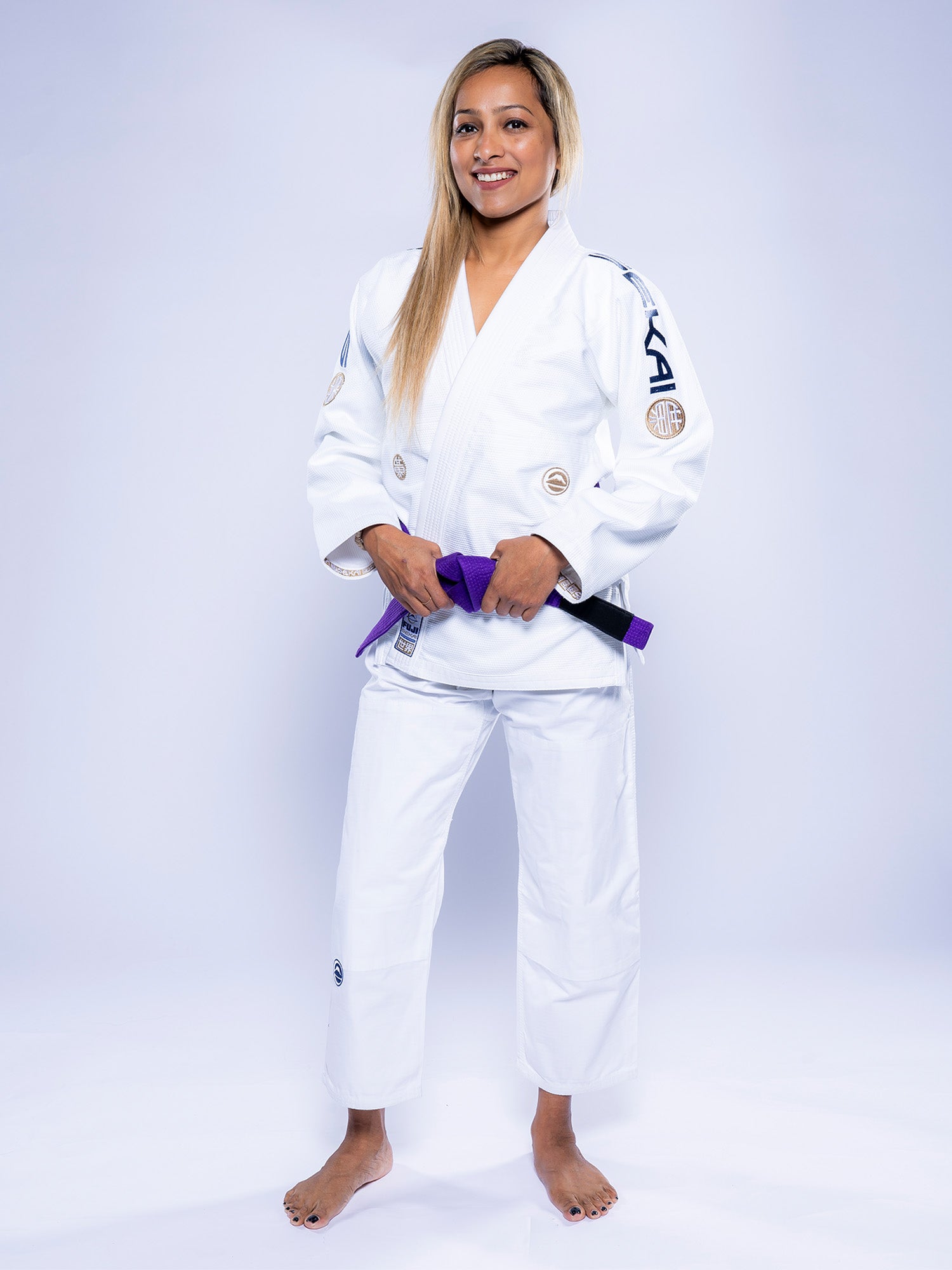 A woman stands barefoot on a white background, smiling and wearing the Womens SEKAI 3.0 BJJ Gi White Navy, with a purple belt tied at her waist.