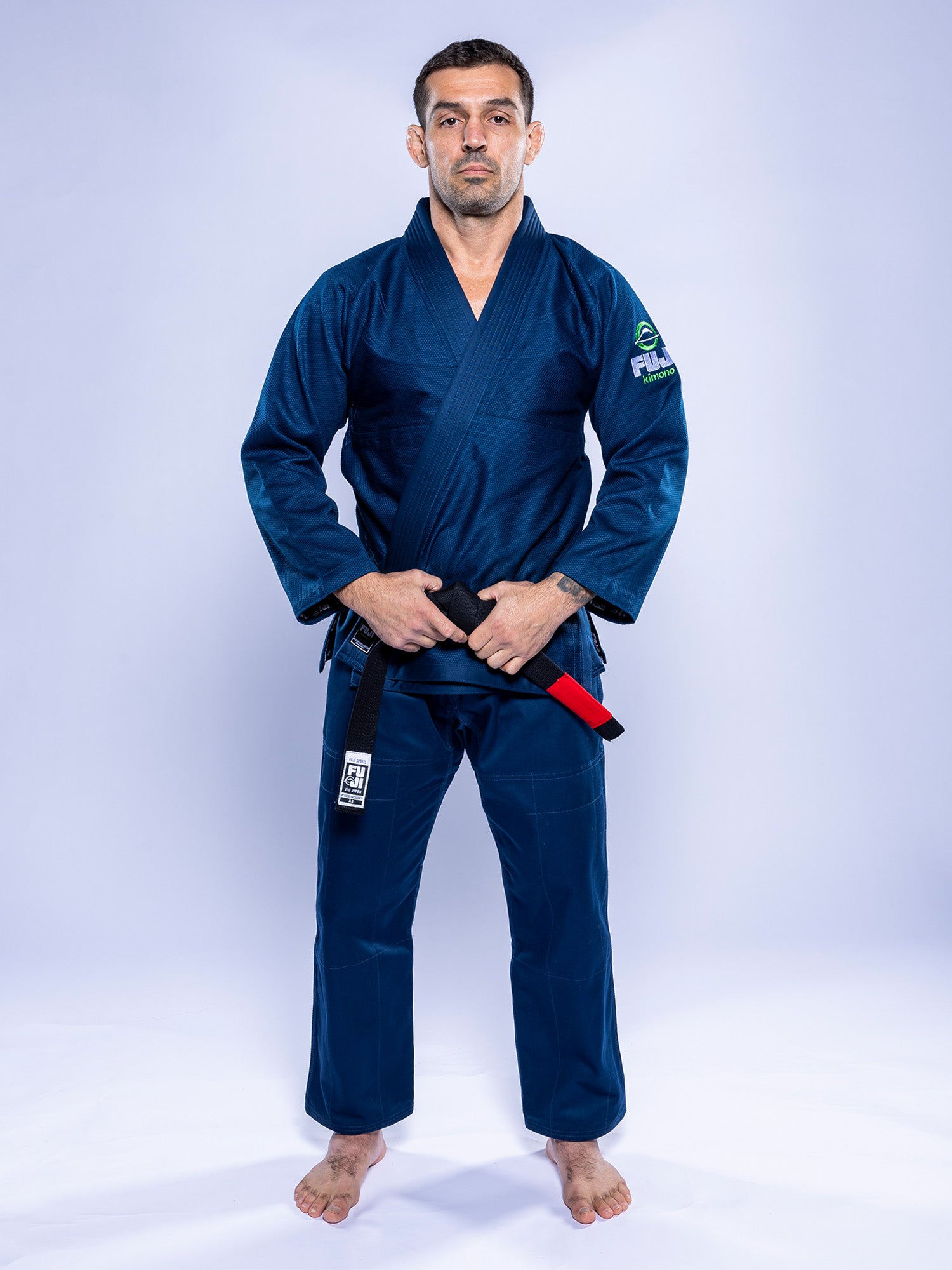 A man in a Lightweight BJJ Gi Navy stands barefoot on a white background, holding a black belt with a red bar in front of him and looking straight ahead with a neutral expression.
