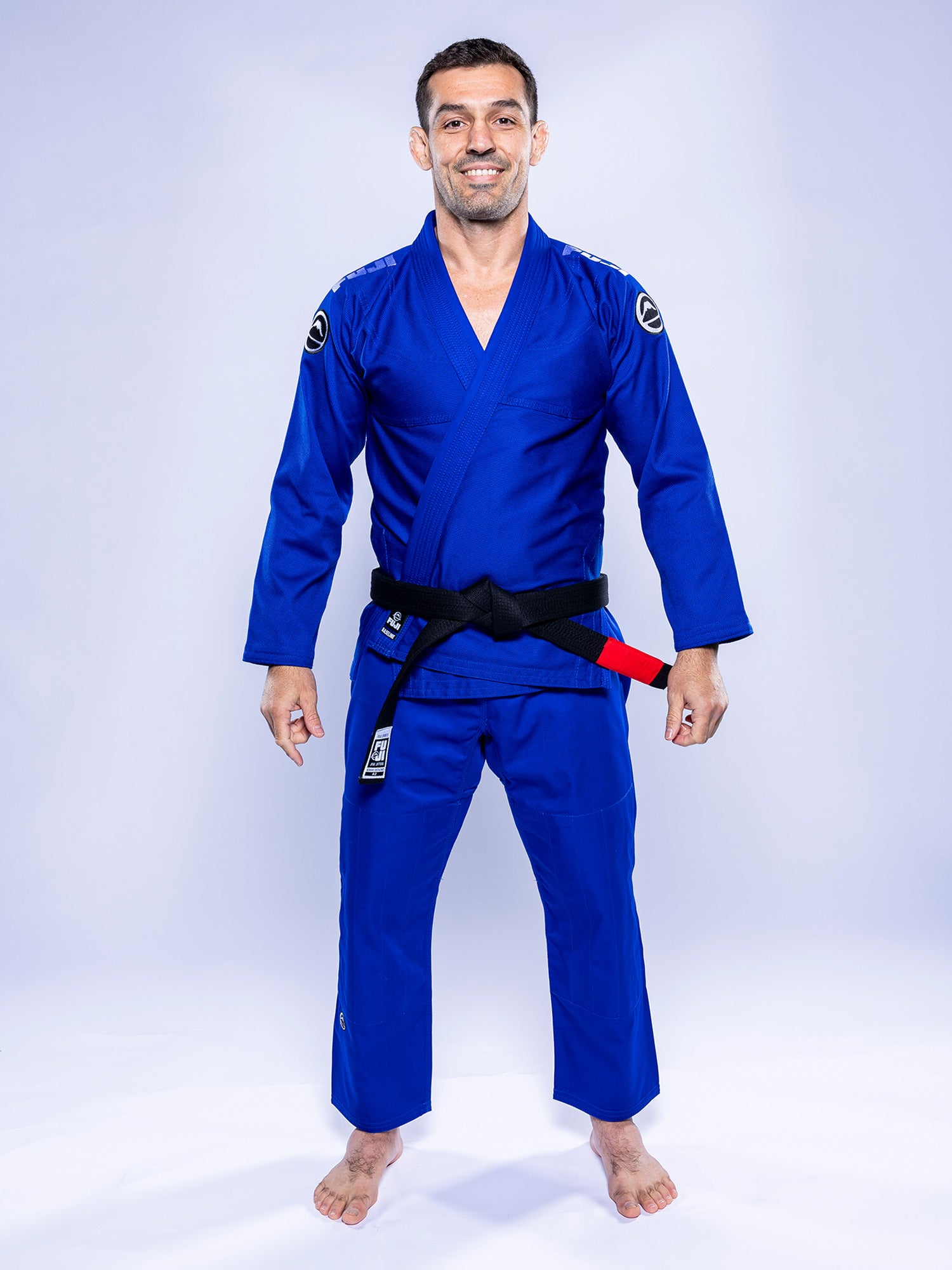 A man wearing the Baseline 2.0 BJJ Gi Blue White and a black belt stands barefoot on a white background, smiling and looking straight ahead.