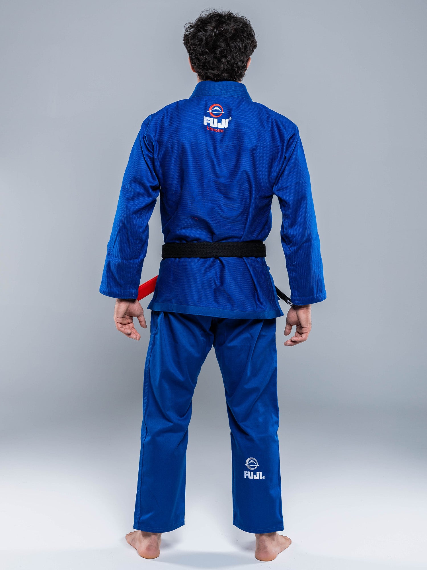 A person stands barefoot facing away, wearing the Lightweight BJJ Gi Blue with a black belt. The FUJI logo is visible on the back of both the jacket and pants. The background is plain gray.