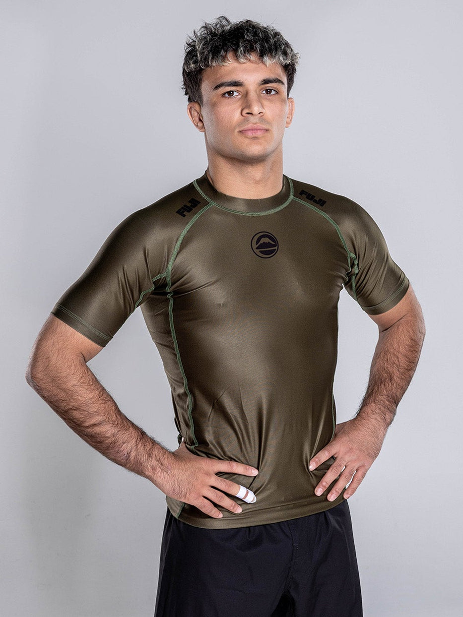 A young man with short curly hair stands barefoot on a white background, wearing the Baseline 2.0 Short Sleeve Rashguard in Military Green and black shorts, hands on his hips, looking confidently at the camera.