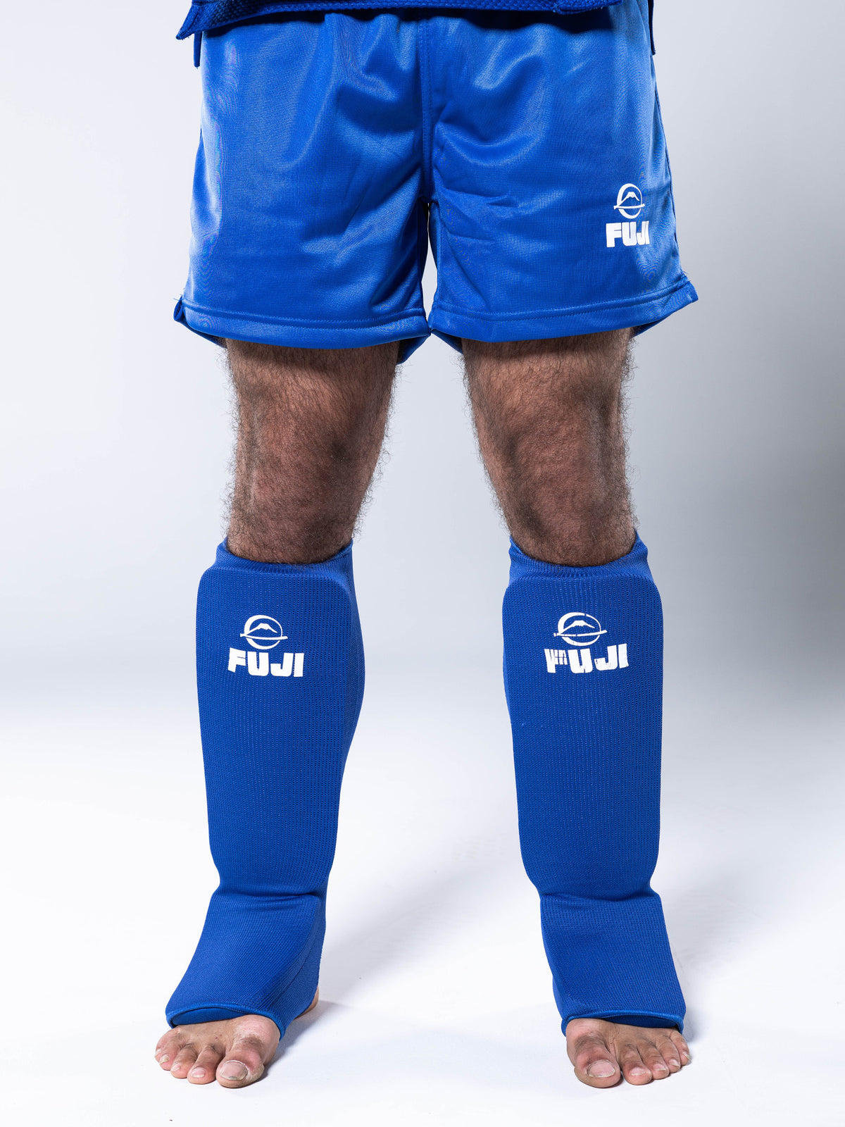 A person stands barefoot wearing blue athletic shorts and FIAS Sambo Combat Shin Guards Blue; only their legs are visible.