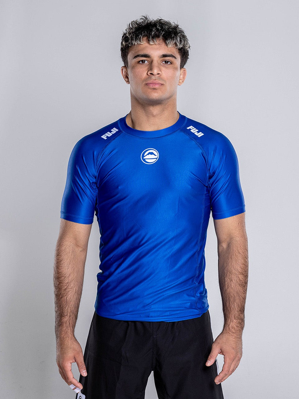 A person with short curly hair stands barefoot on a white background, facing forward with arms relaxed, wearing the Baseline 2.0 Short Sleeve Rashguard Navy and black shorts.