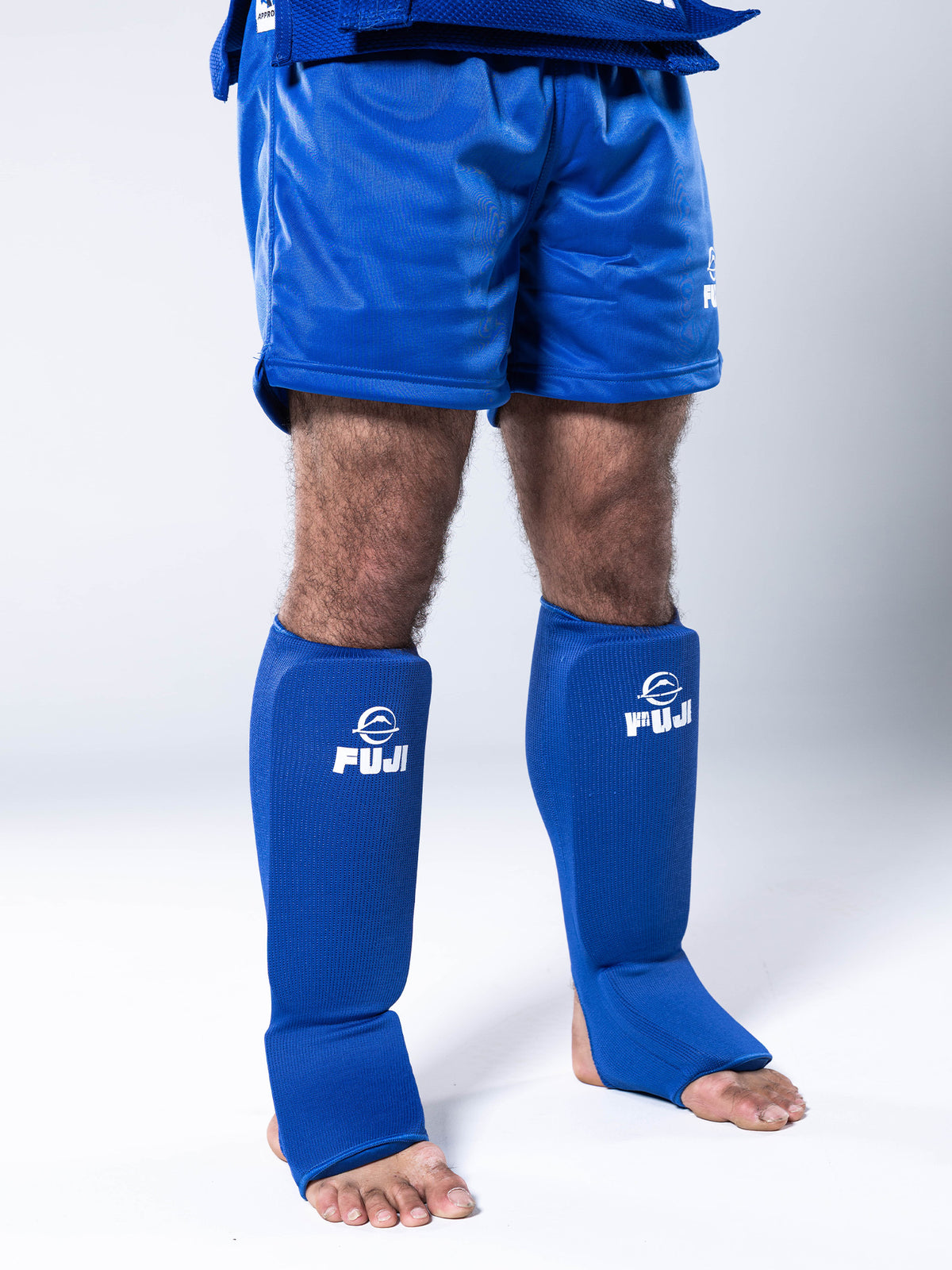 A person stands barefoot on a white background, wearing FIAS Sambo Combat Shin Guards Blue and blue martial arts shorts. The FUJI logo appears in white on the shin guards, with only their legs and lower torso visible.
