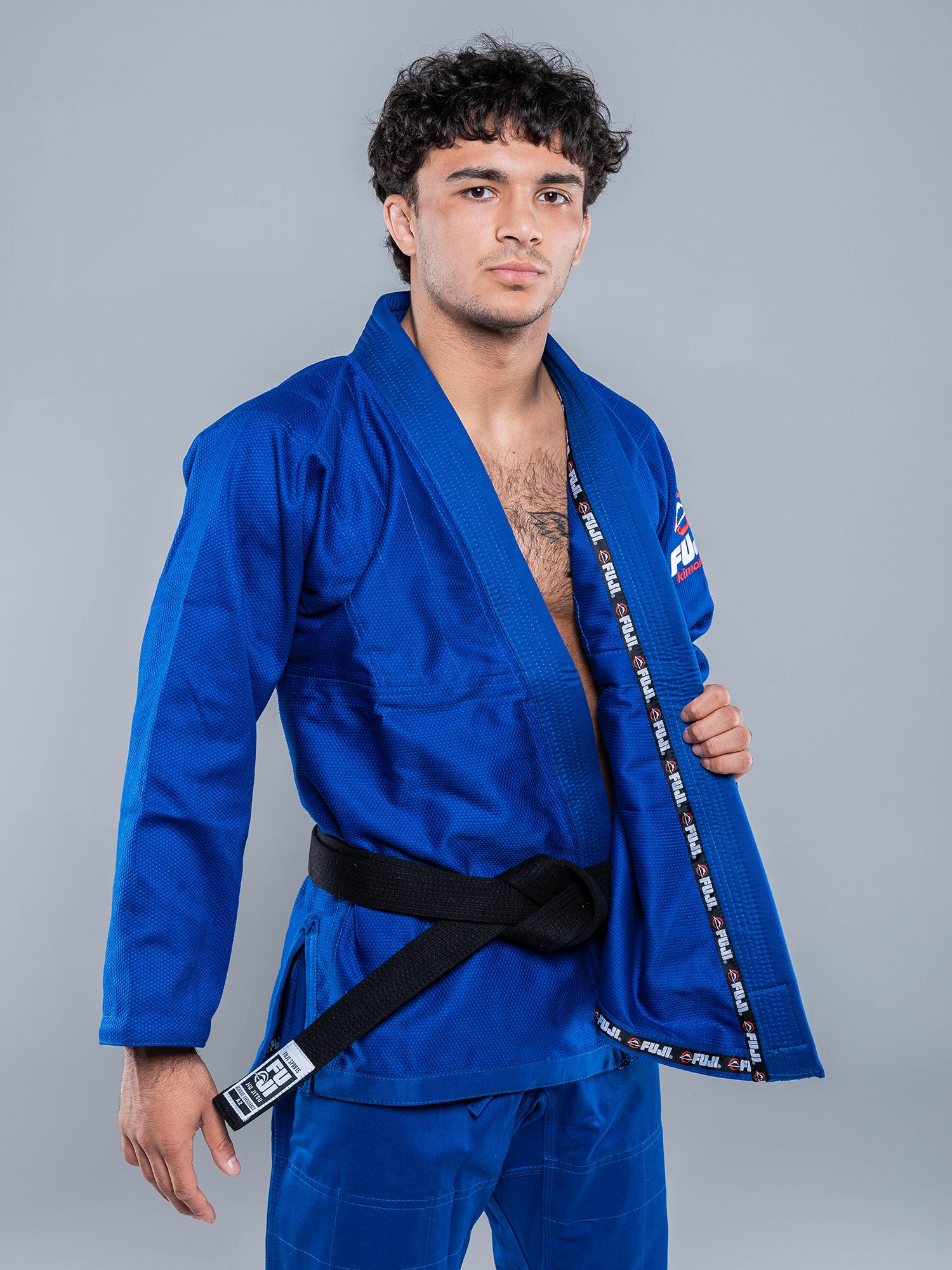 A young man with short curly hair wears the Lightweight BJJ Gi Blue and a black belt, standing confidently against a plain gray background while holding one side of his open jacket.