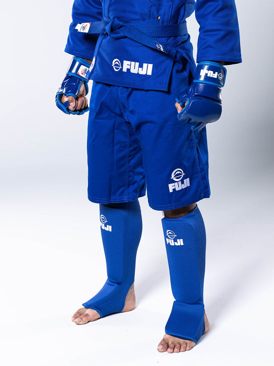 A person wearing a blue martial arts uniform, including FIAS Combat Sambo Shorts Blue, a jacket, shin guards, and fingerless gloves.
