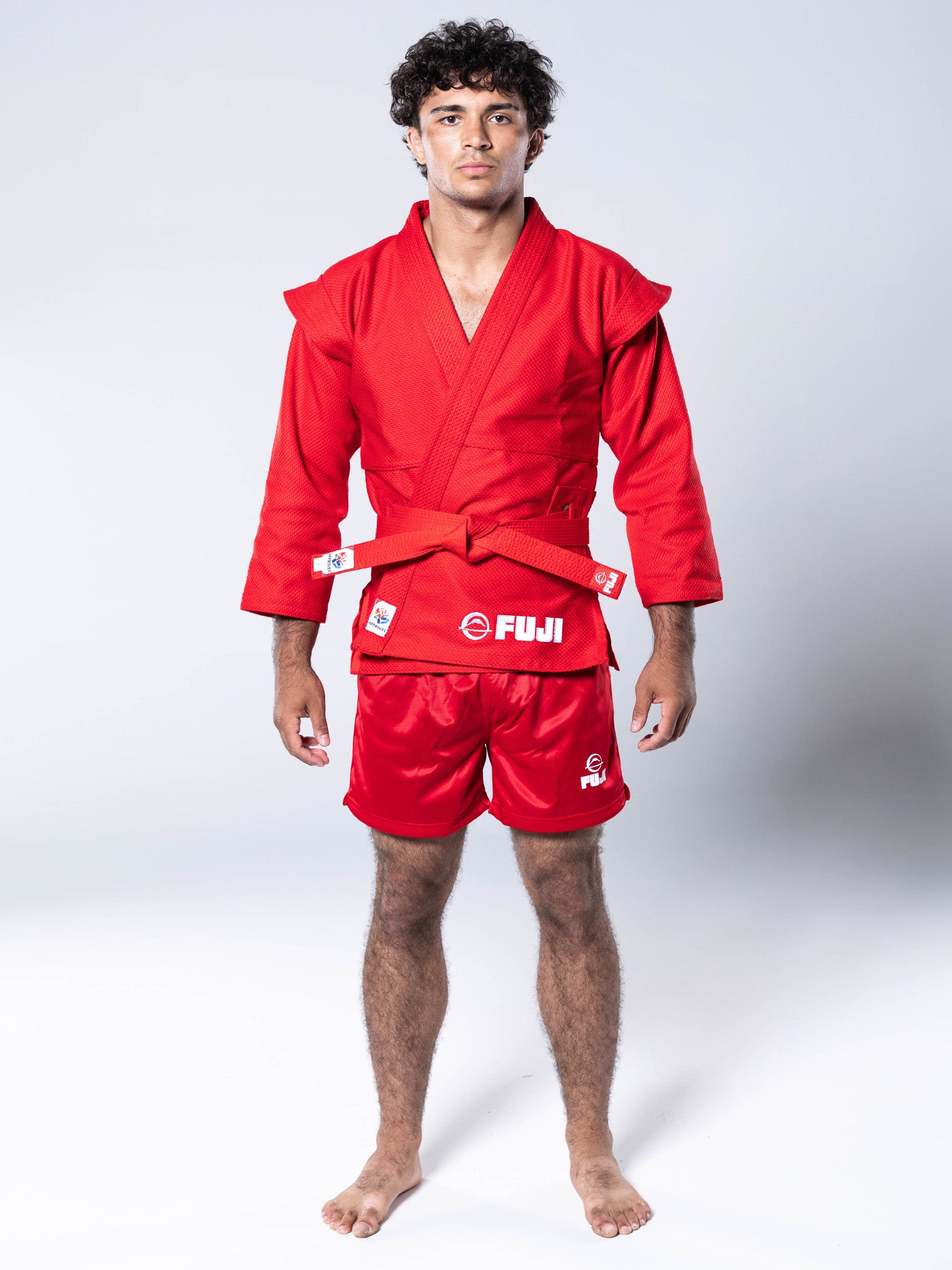 A young man stands barefoot on a white background, wearing the FIAS Sambo Kurtka Jacket Red with matching red shorts and a red belt, both featuring FUJI branding, as he looks forward with a neutral expression.