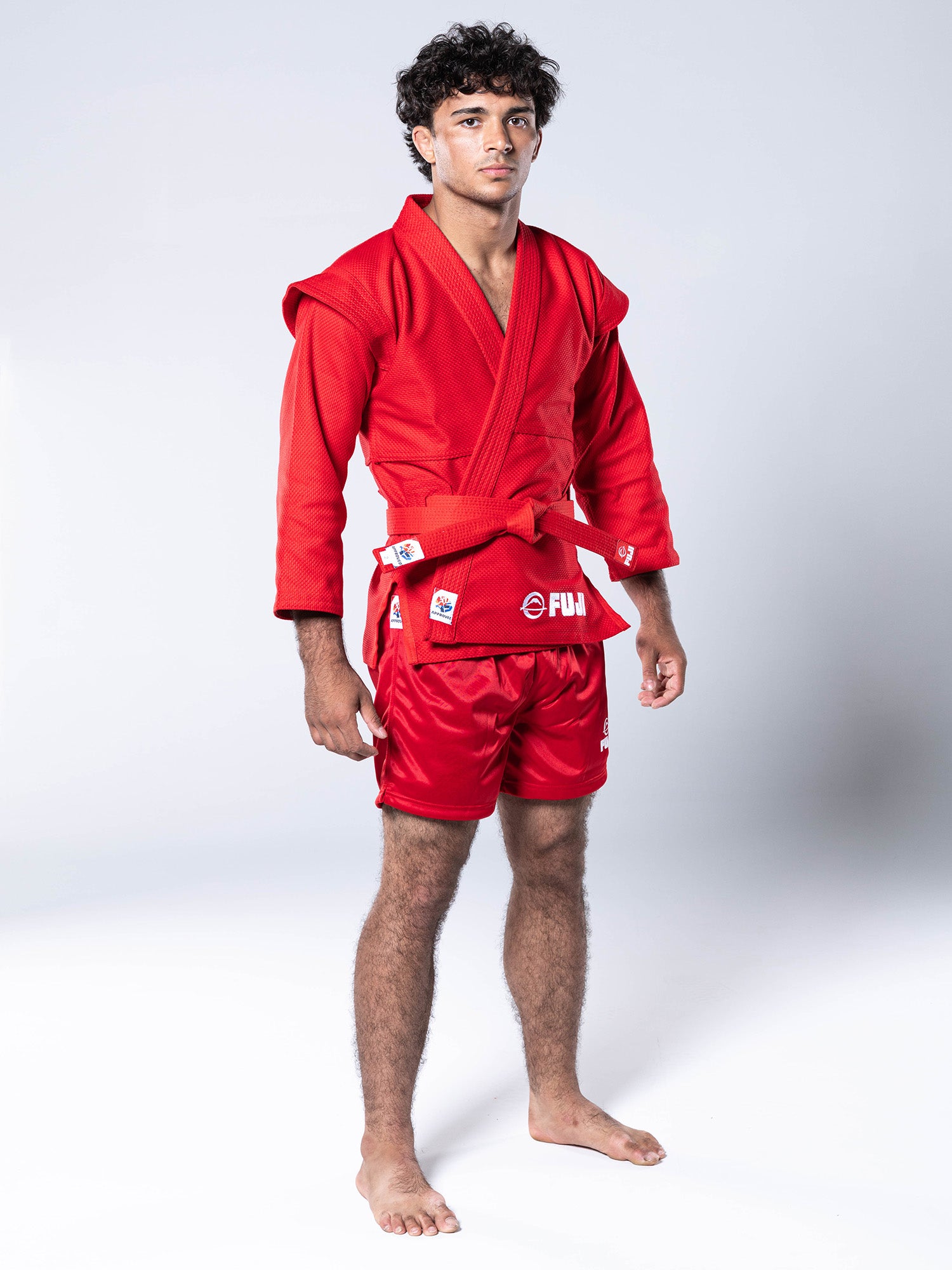 A young man stands barefoot in a red FIAS Sambo Kurtka Jacket with a matching red belt and shorts, against a plain light background. He looks forward with a neutral expression.