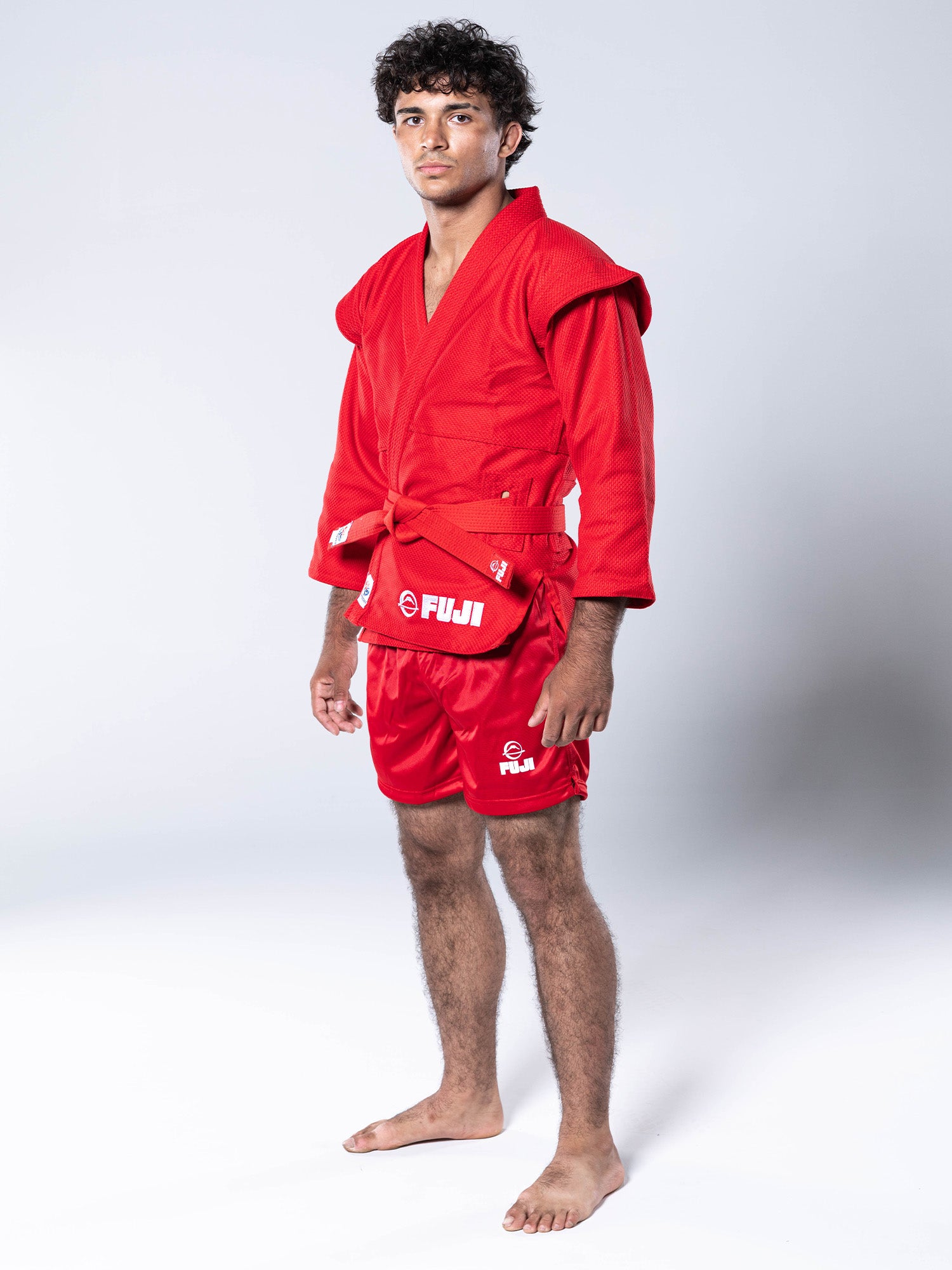A man wearing a FIAS Sambo Kurtka Jacket Red with a matching red belt, paired with red shorts, stands barefoot against a white background.