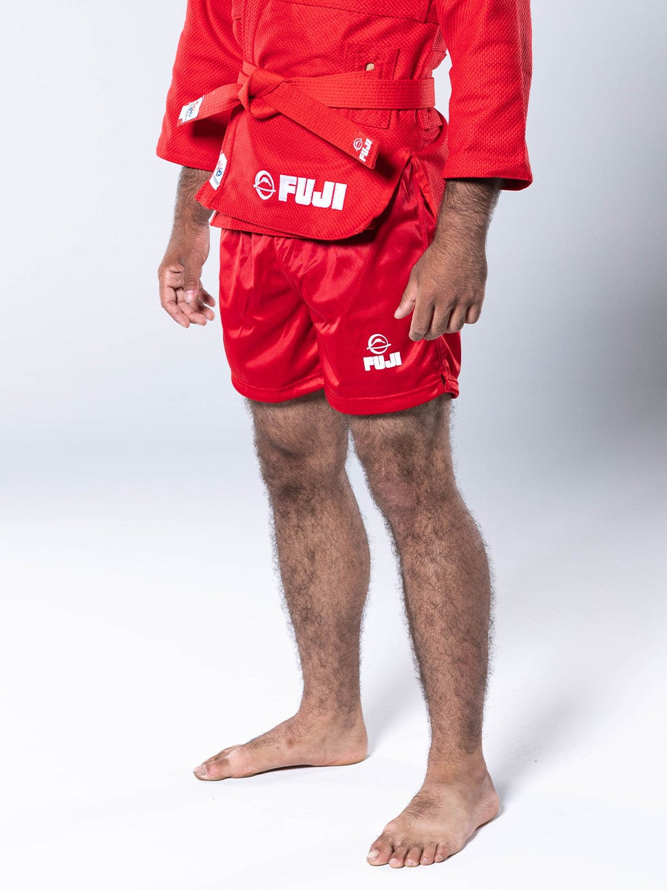 A young man stands barefoot in FIAS Sambo Shorts Red, paired with a red martial arts gi top, against a plain white background.