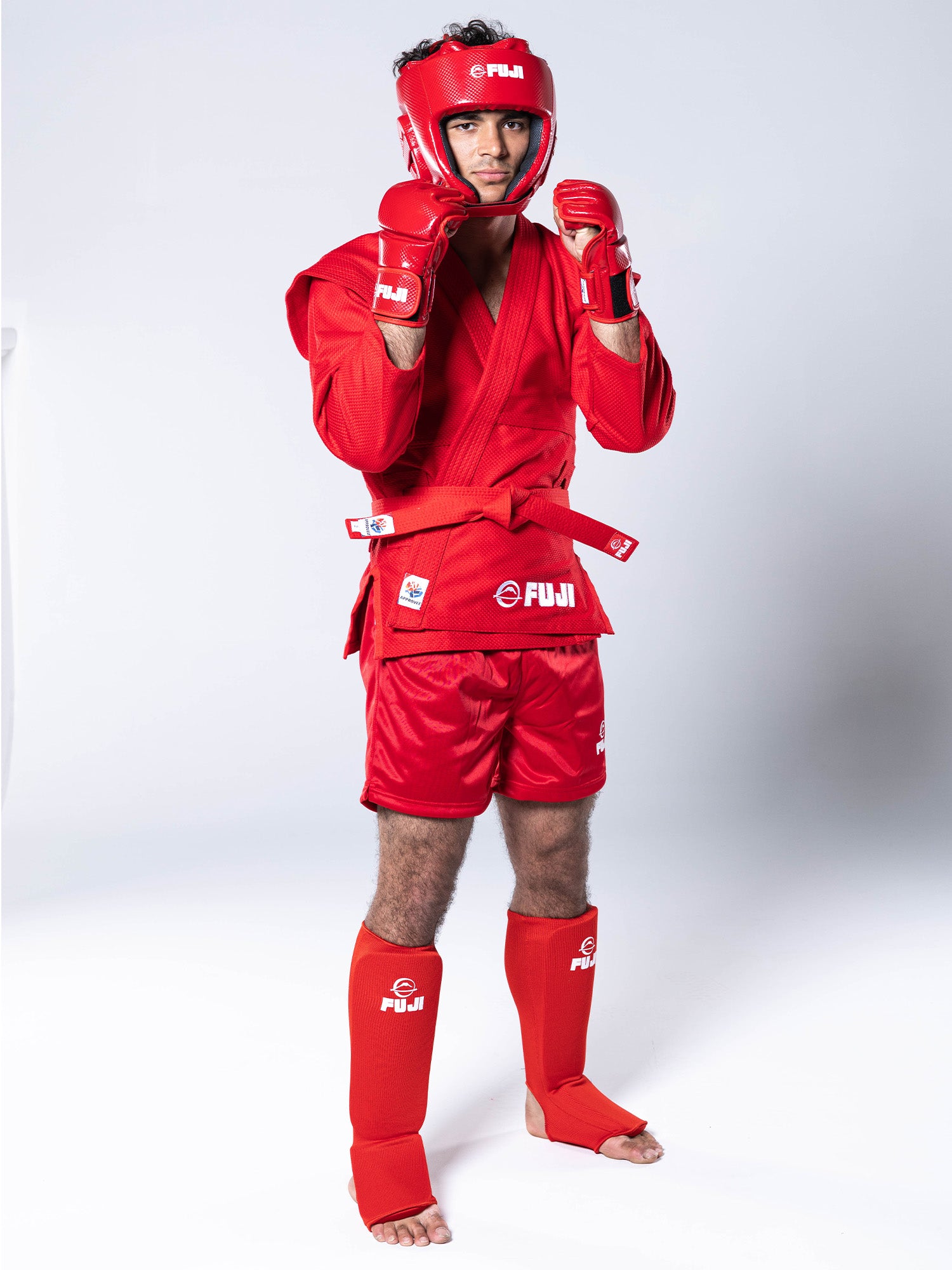 Male martial artist in a fighting stance wearing the FIAS Sambo Combat Headgear Red along with matching red gloves, jacket, shorts, and shin guards.