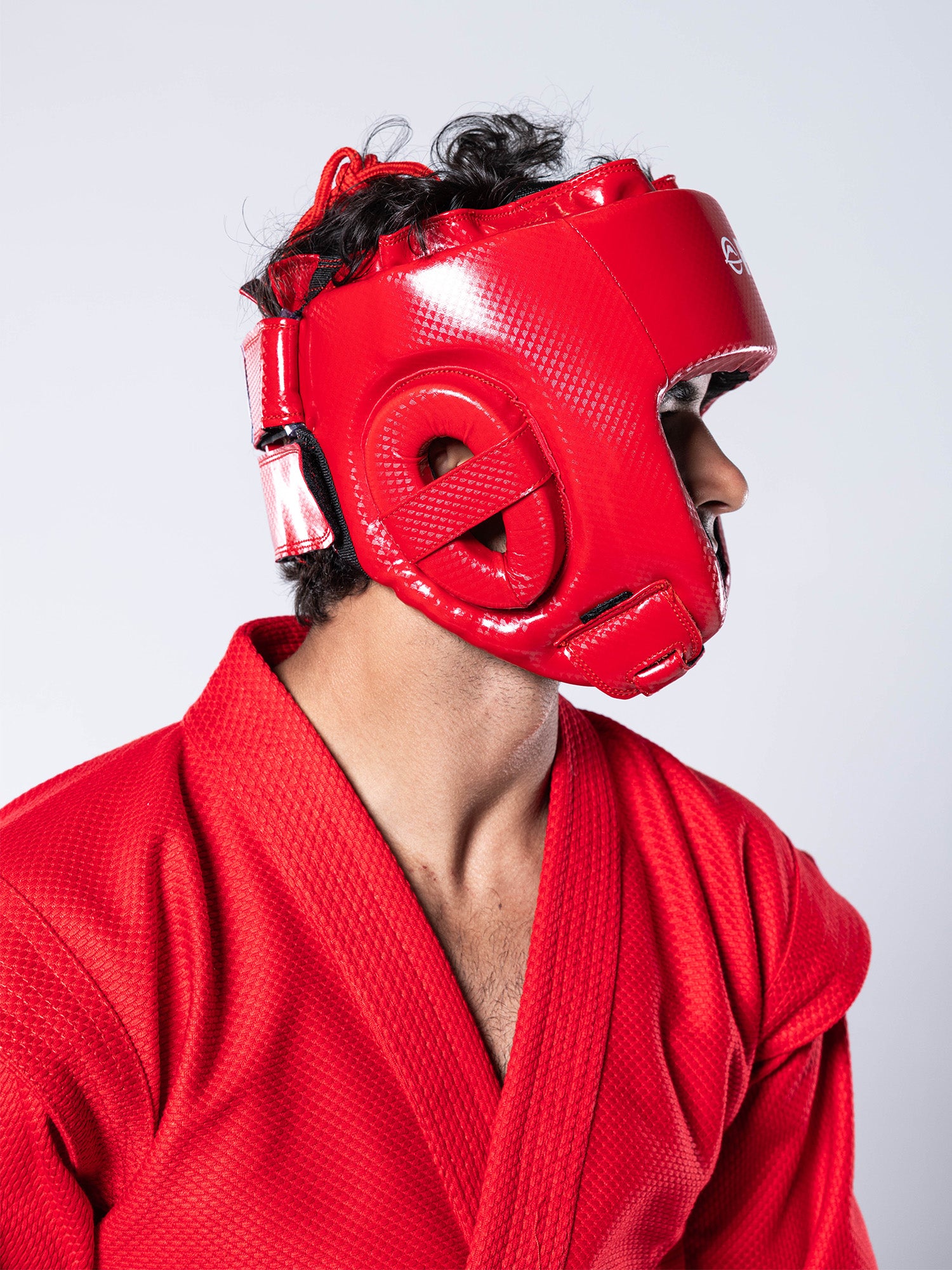 A person wearing the FIAS Sambo Combat Headgear Red and a red martial arts uniform looks to the side against a plain light background.