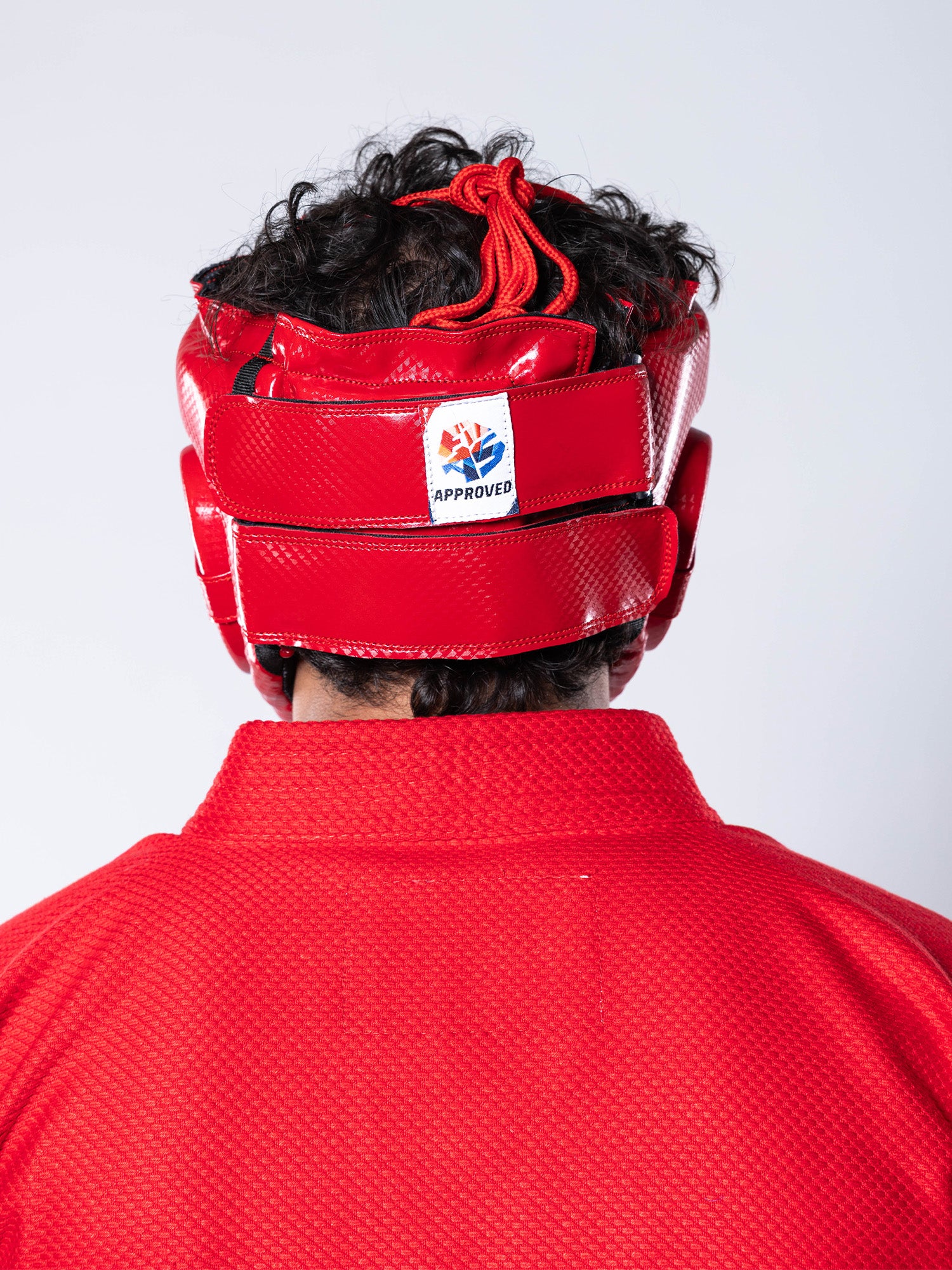 A person wearing a red martial arts uniform and the FIAS Sambo Combat Headgear Red with an approval label, shown from the back against a plain background.