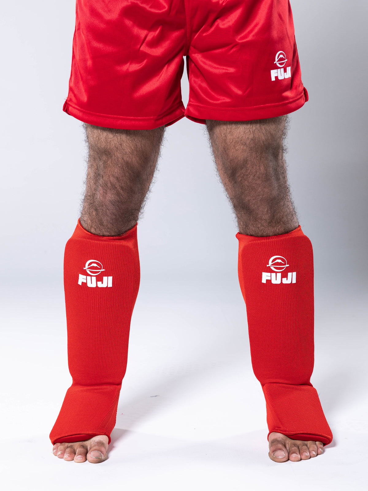 A person wearing red shorts and FIAS Sambo Combat Shin Guards Red, standing barefoot on a white background.