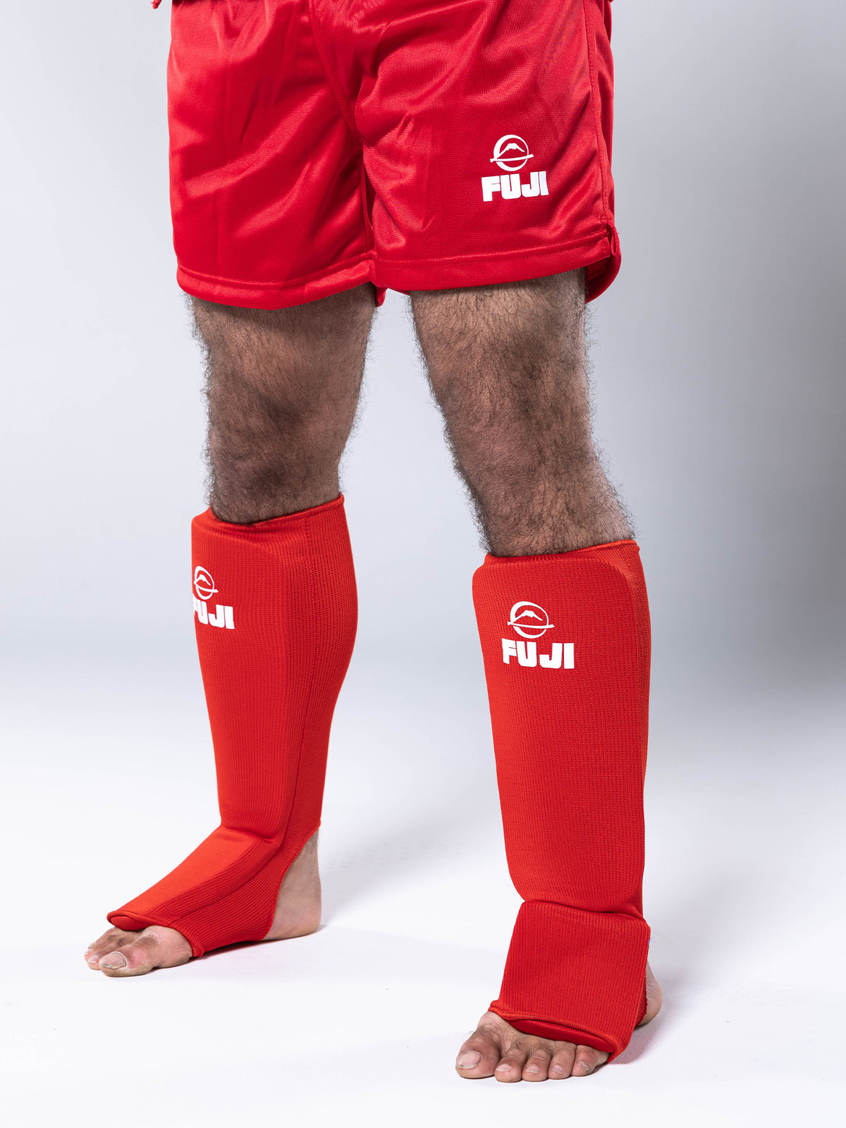 A person wearing the FIAS Sambo Combat Shin Guards Red, along with red shorts, standing barefoot against a light background. Only the legs and lower torso are visible.