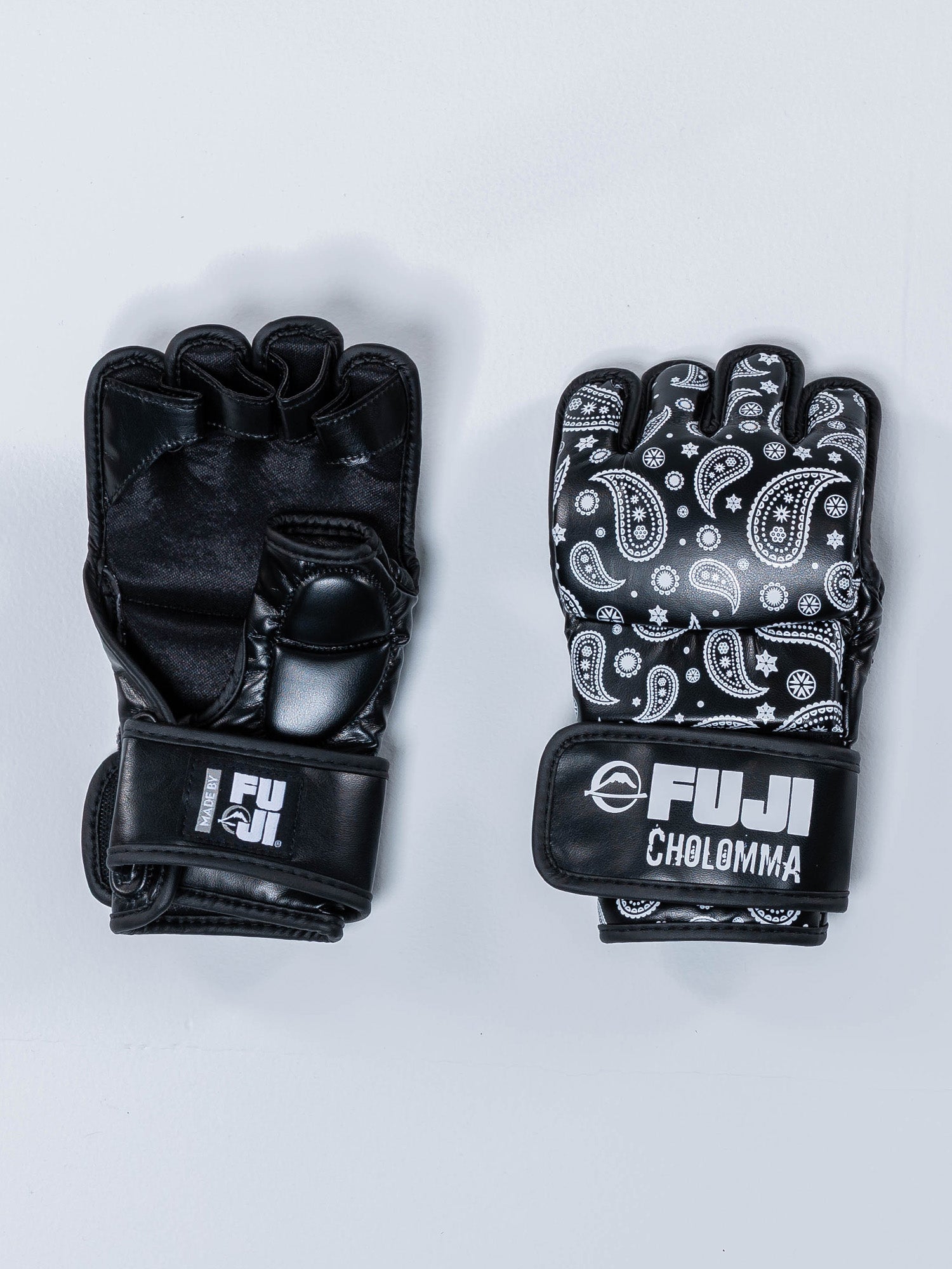 Two black CholoMMA Paisley MMA Gloves are displayed on a light surface. The right glove has a white paisley pattern and FUJI CHOLOMMA branding, while the left is solid black with FUJI branding on the wrist strap.
