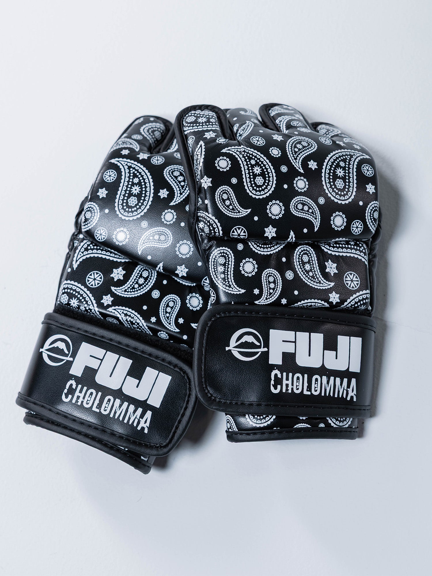 A pair of CholoMMA Paisley MMA Gloves in black with white paisley patterns and stars, featuring bold FUJI and CHOLOMMA branding on the wrist straps, resting on a light surface.