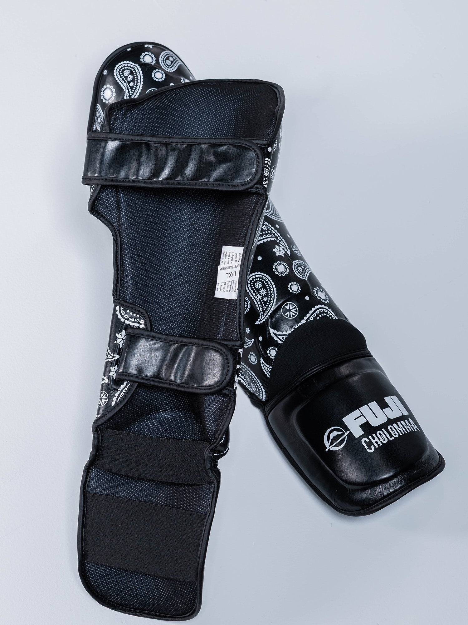 CholoMMA Paisley Shin Guards in black feature white paisley patterns and "FUJI CHLOHOMA" on the front, shown on a light gray surface.